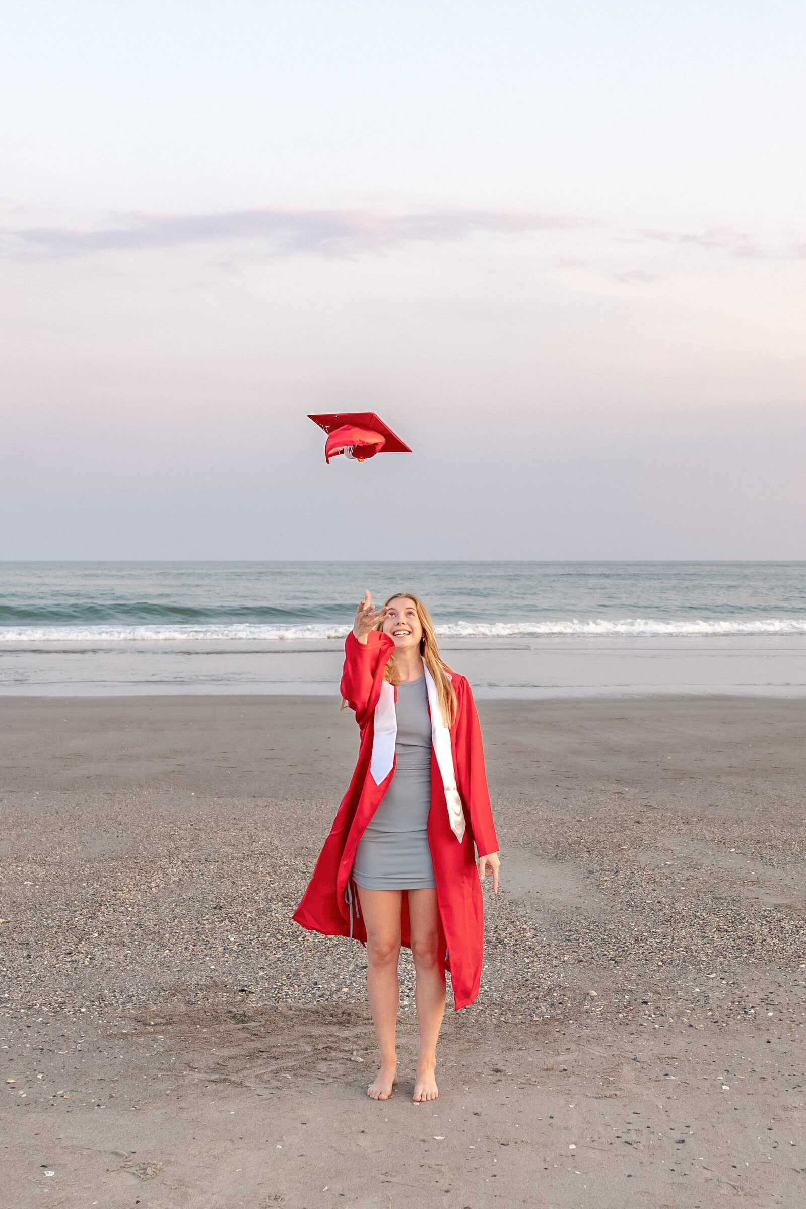 A college senior in a red gown joyfully tossing her graduation cap in the air on the beach with waves in the background in North Topsail Beach, North Carolina. This energetic image captures the celebration of graduation, perfect for memorable senior moments by the sea.