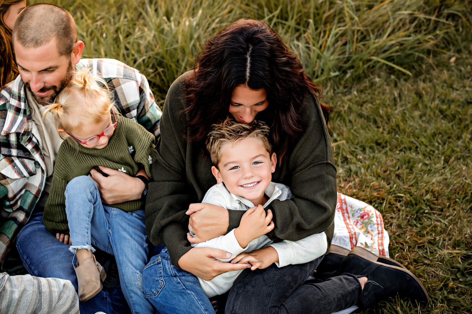 A family sitting on grass in a warm embrace captures the essence of outdoor portraits. A woman hugs a smiling young boy while a man and a girl with red glasses and braids sit close, all wearing casual clothes. The scene conveys warmth and joy, reminiscent of a Bloomington family photographer’s perfect shot.