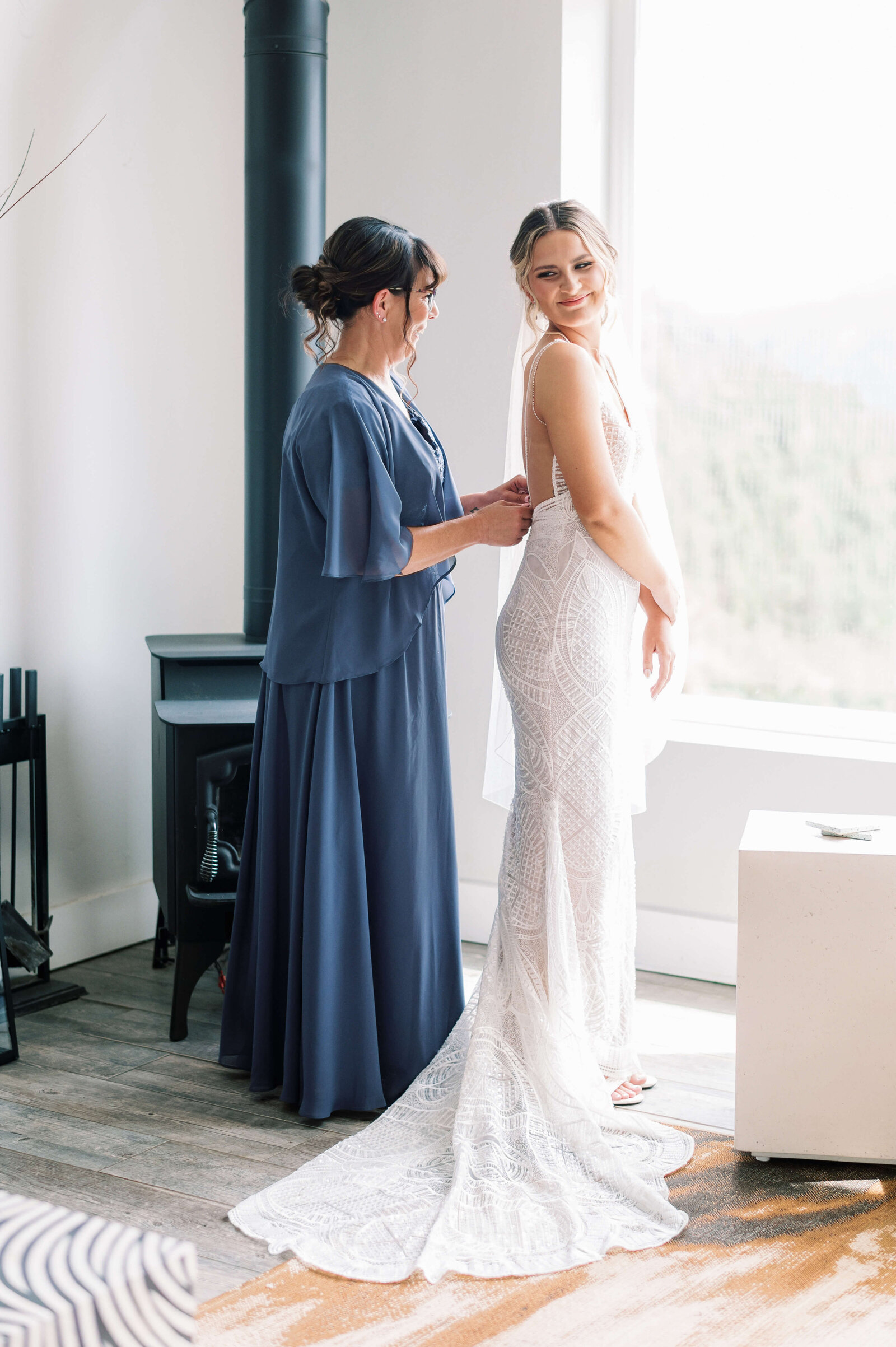 Mother of the bride, wearing a long blue dress, zips up the gown of the bride