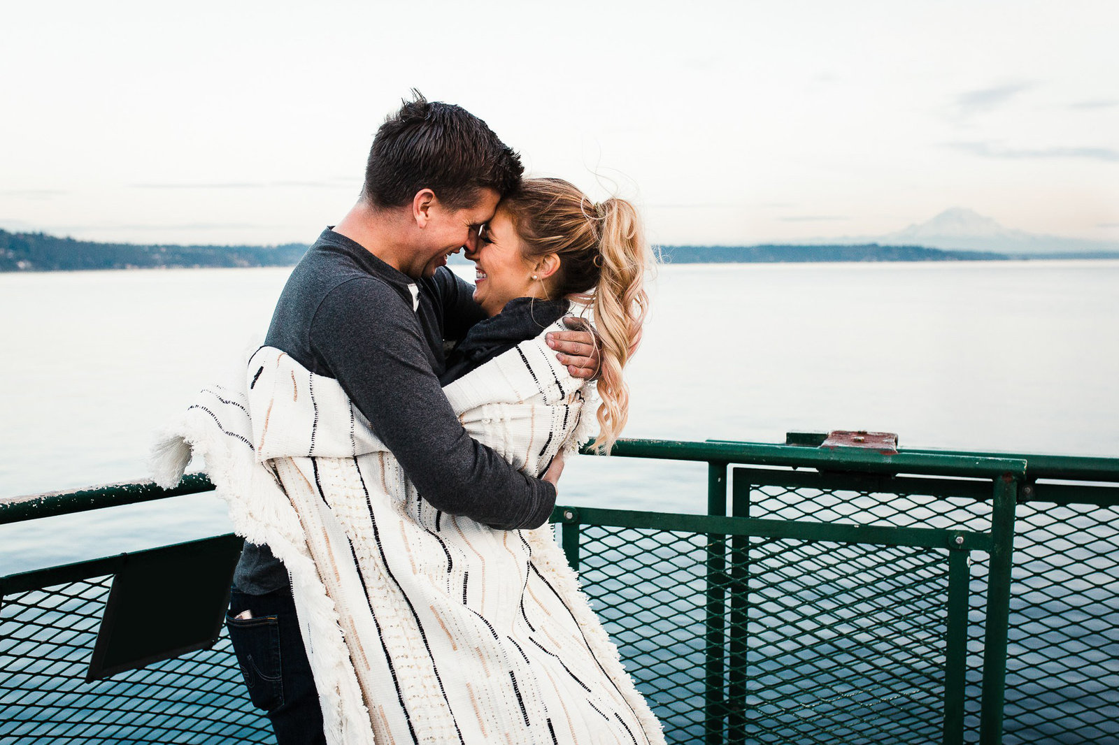 a couple snuggles under a blanket as they ride the ferry across puget sound and enjoy views of mt rainier in the background during their adventure engagement session with amy galbraith