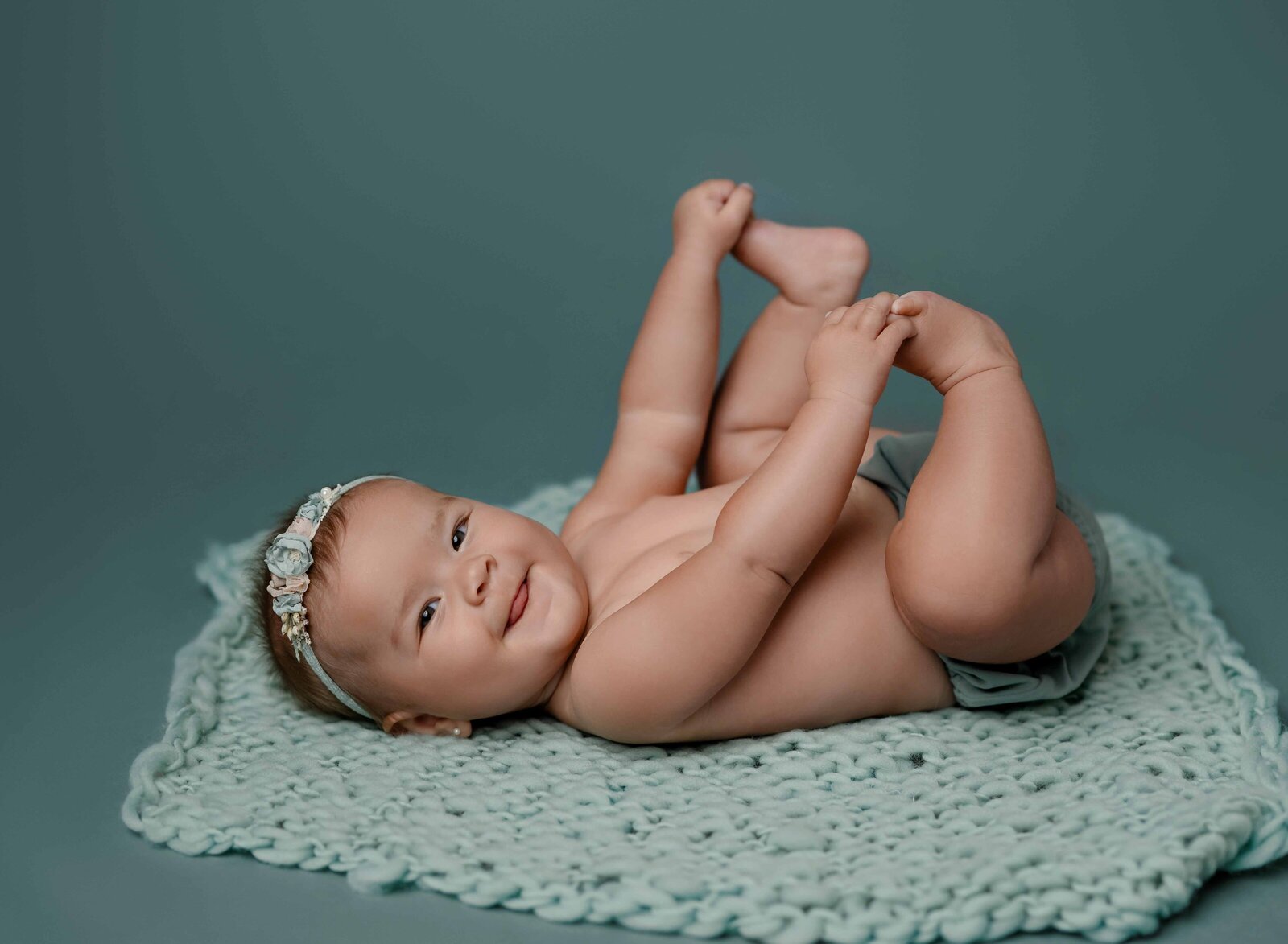 Baby girl in a teal diaper cover, grabbing her toes and smiling.