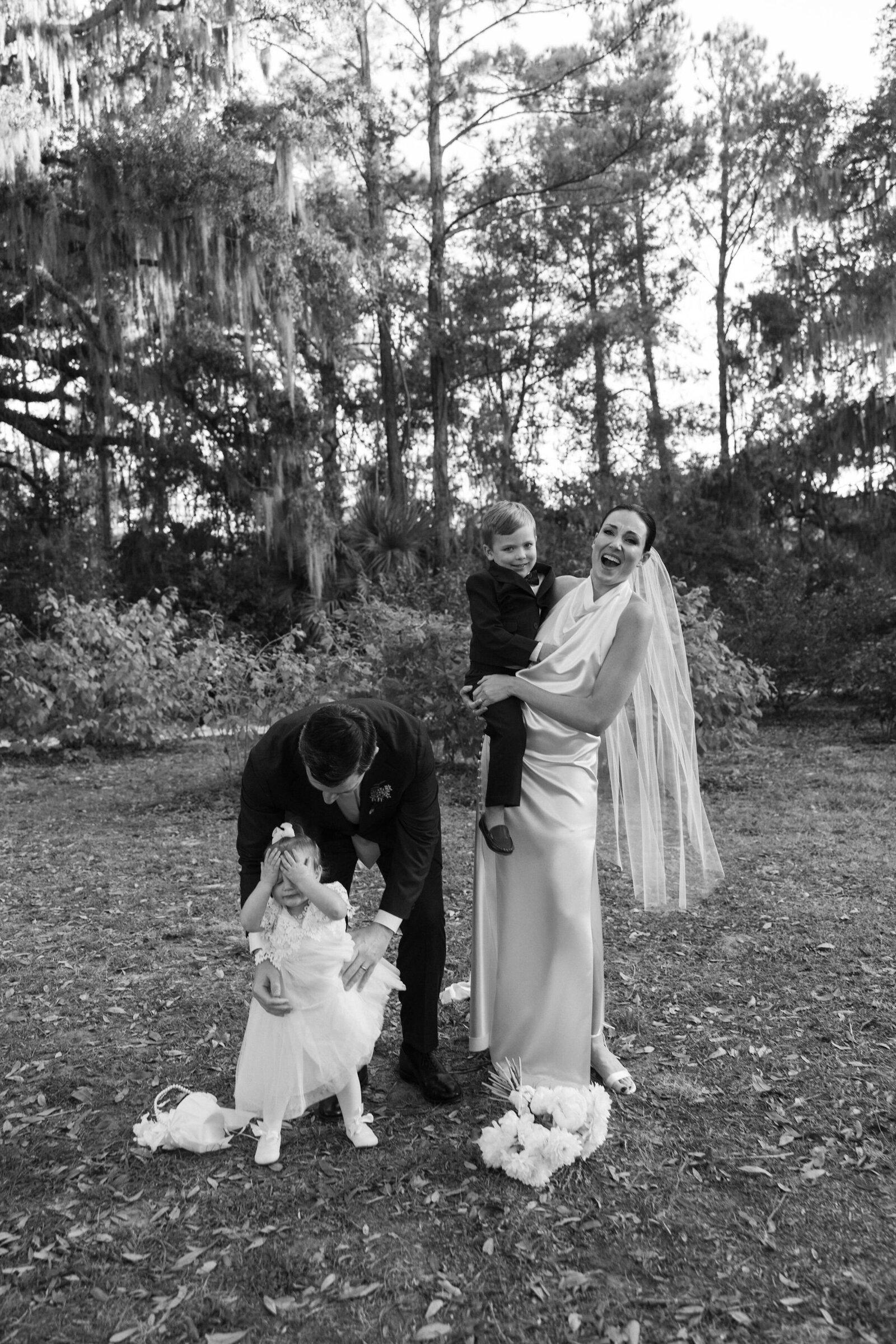 Candid black & white family wedding photo with bride holding ring bearer and groom picking up crying flower girl 