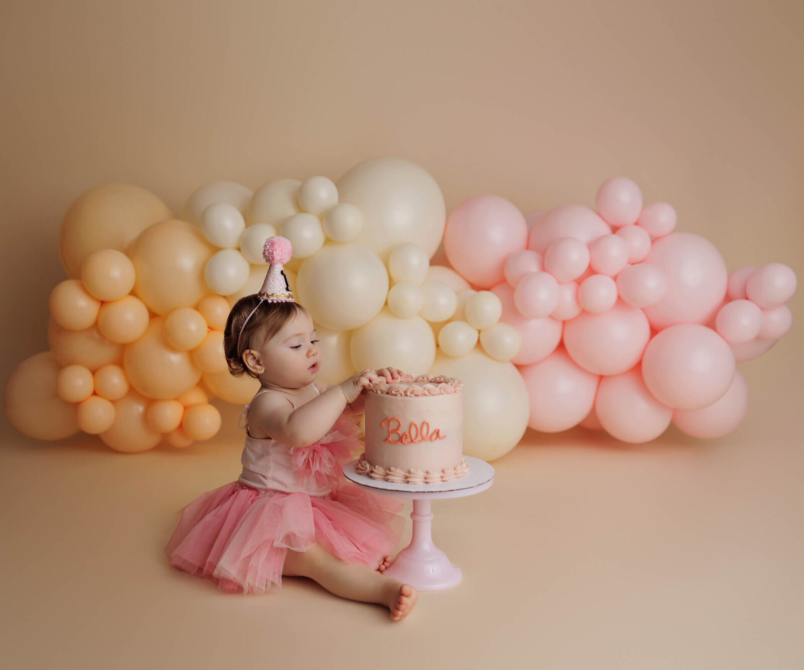 A sweet toddler in a pink tutu dress and a matching party hat sits on a beige backdrop with a festive balloon garland in soft shades of peach, cream, and pink. The child is gently touching a pink frosted cake on a white cake stand, with the name "Bella" written on it, celebrating a special occasion.