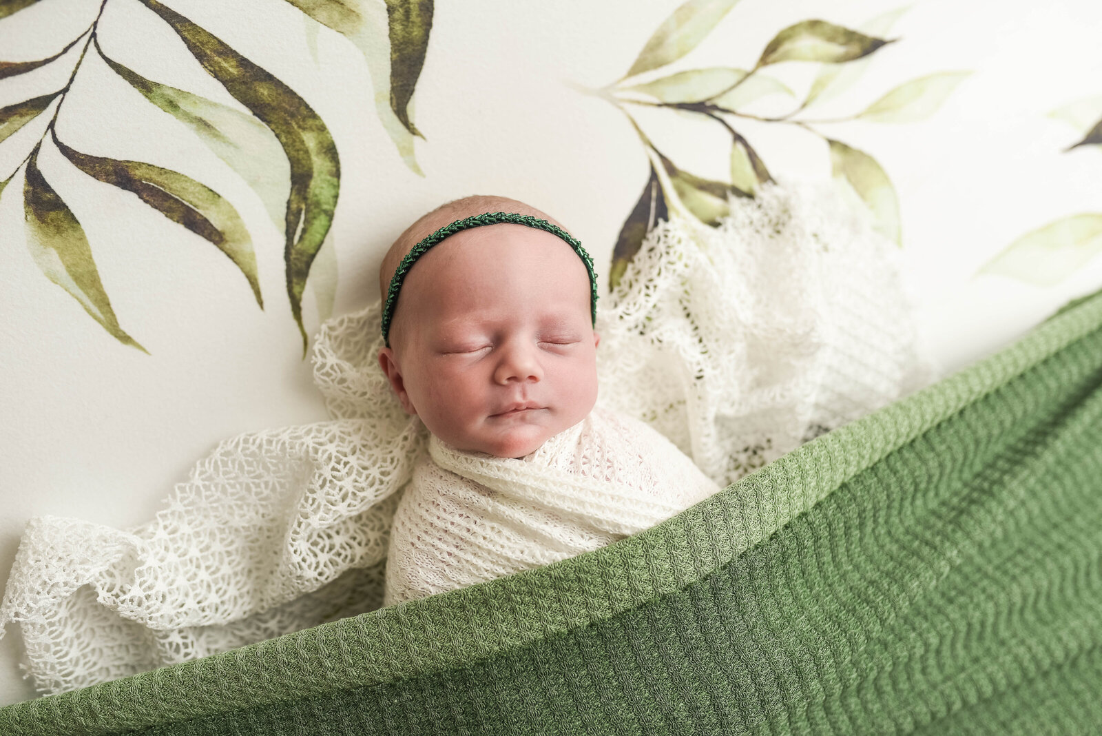 new born baby laying  in white blanket with green plants wrapped in a green blanket