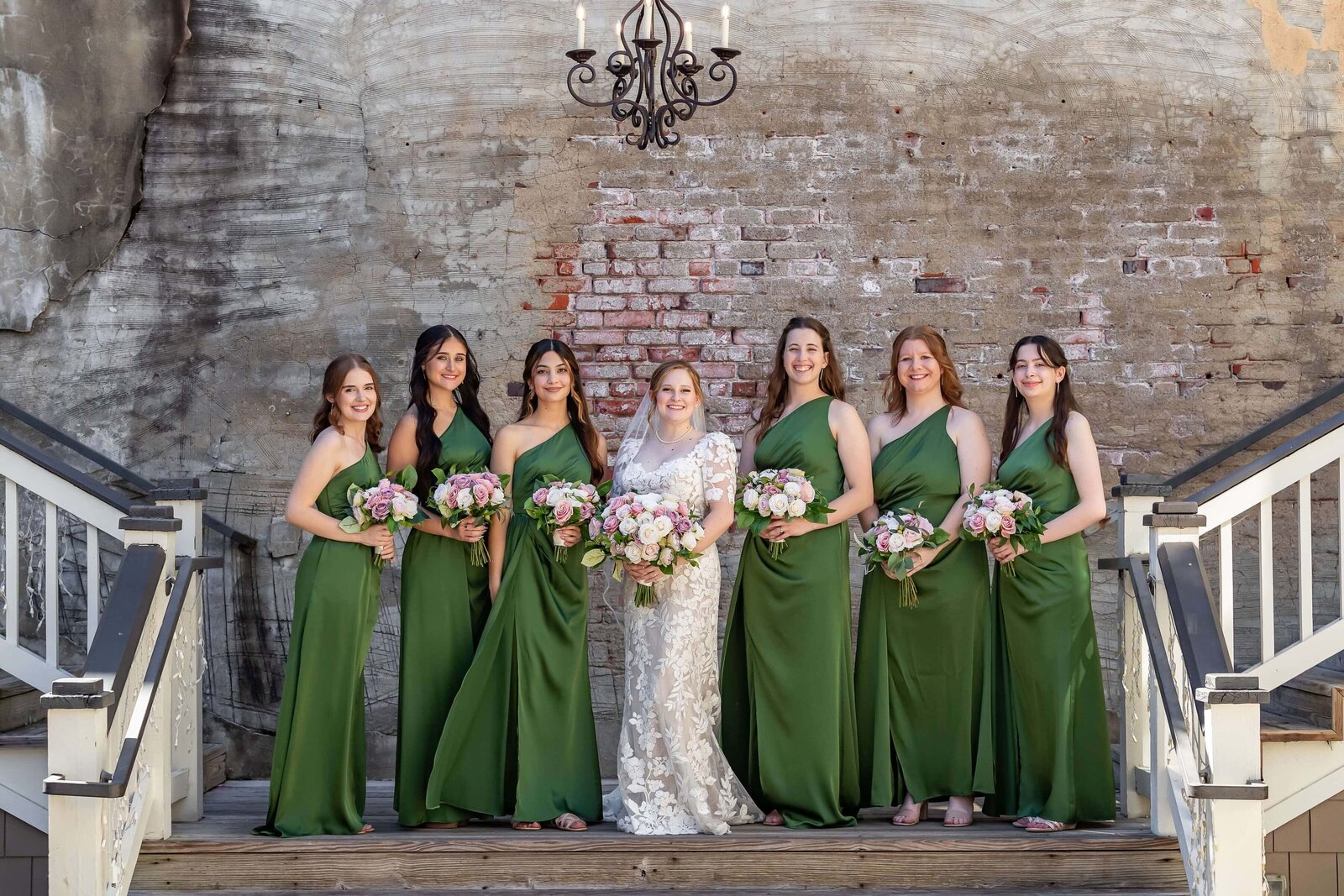 A bride in a white lace gown stands with six bridesmaids in matching green dresses, all holding pink and white bouquets, posing on outdoor wooden stairs against a rustic brick and plaster wall.