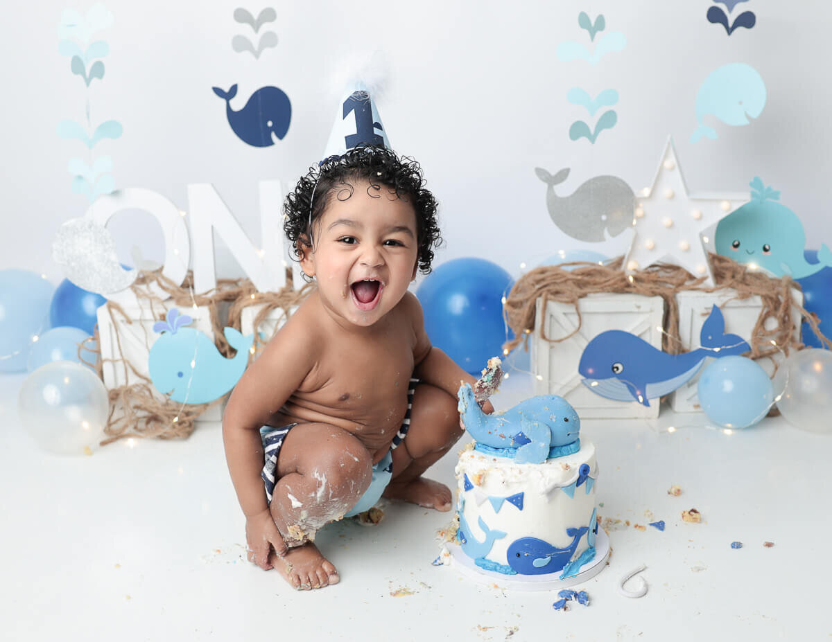 A playful baby covered in frosting during a whale-themed cake smash photo shoot in a cozy studio setting.
