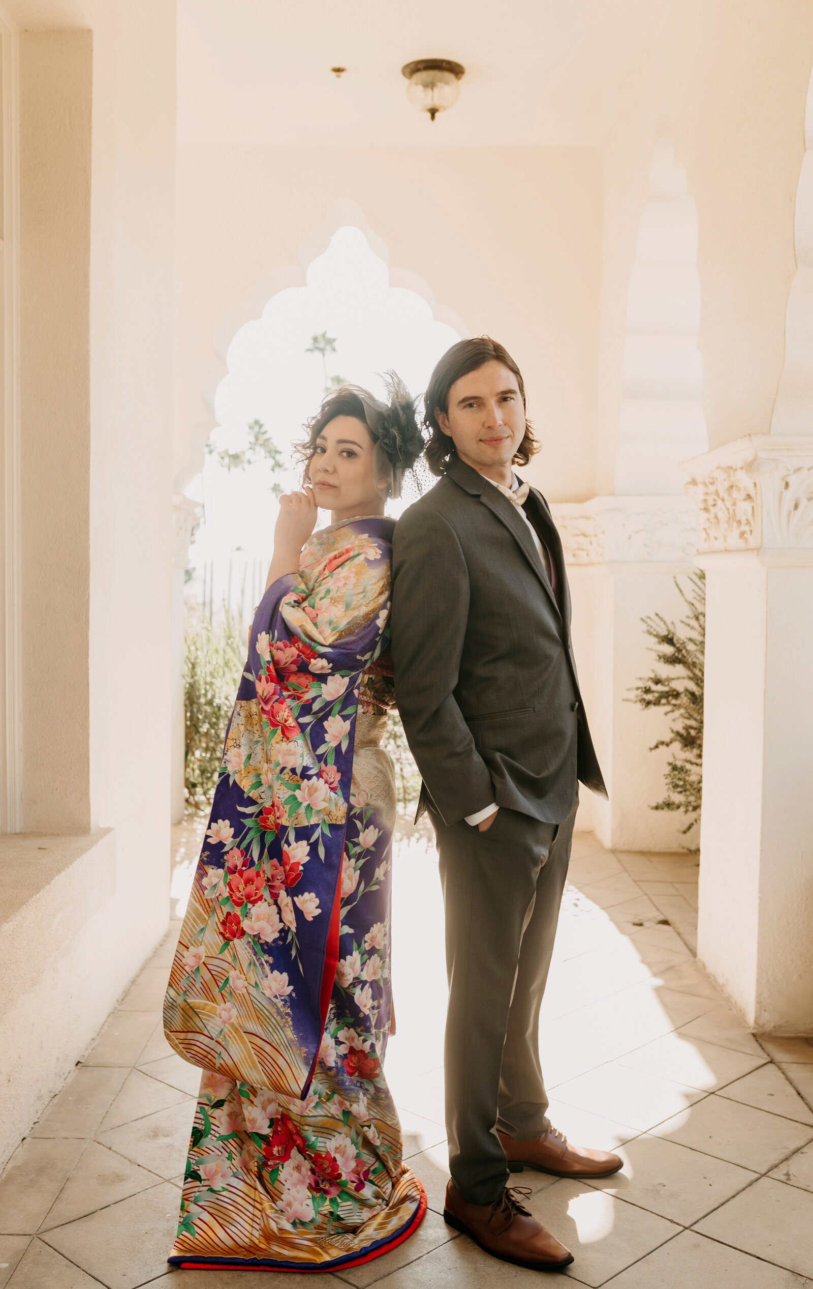 Bride in elegant purple floral wedding kimono rental standing back-to-back with groom in charcoal suit under the white arches at Brand Park, Los Angeles California | ブランドパーク白いアーチ下で紫色振袖の花嫁とスーツ姿の新郎が背中合わせに立つ着物レンタルウェディング