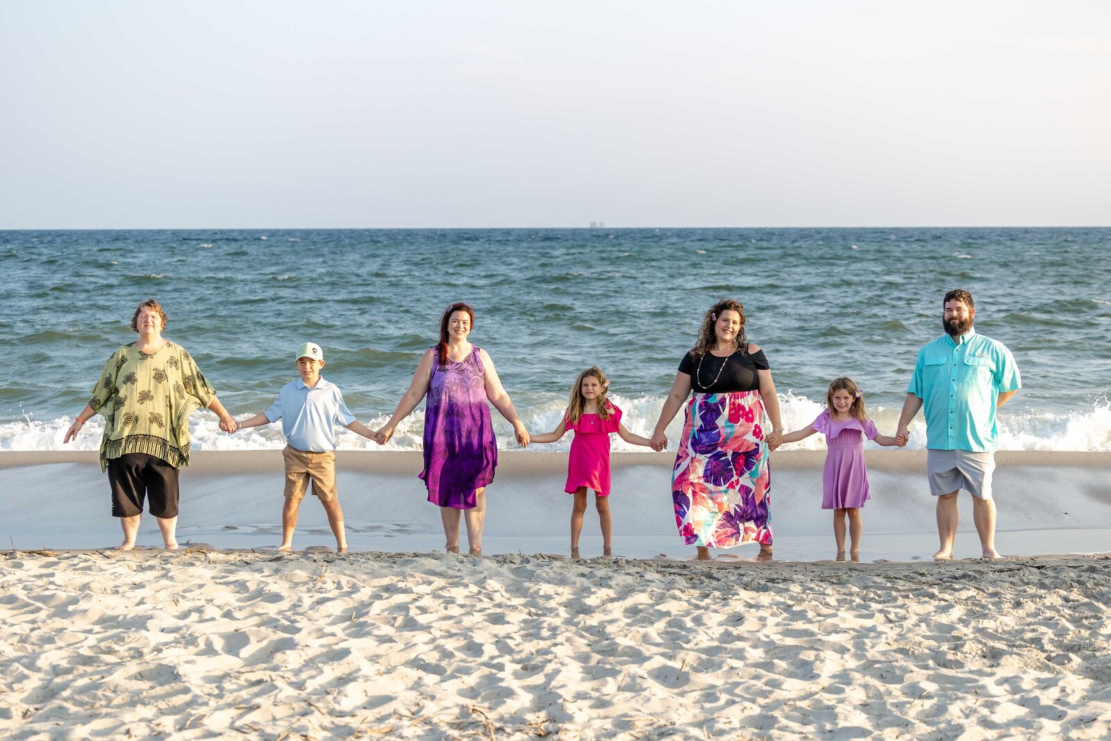 A family holds hands on the sandy shore, dressed in vibrant and colorful attire. The ocean waves and bright sky create a cheerful atmosphere, perfect for destination family photographer NC, capturing unity and joy by the sea.