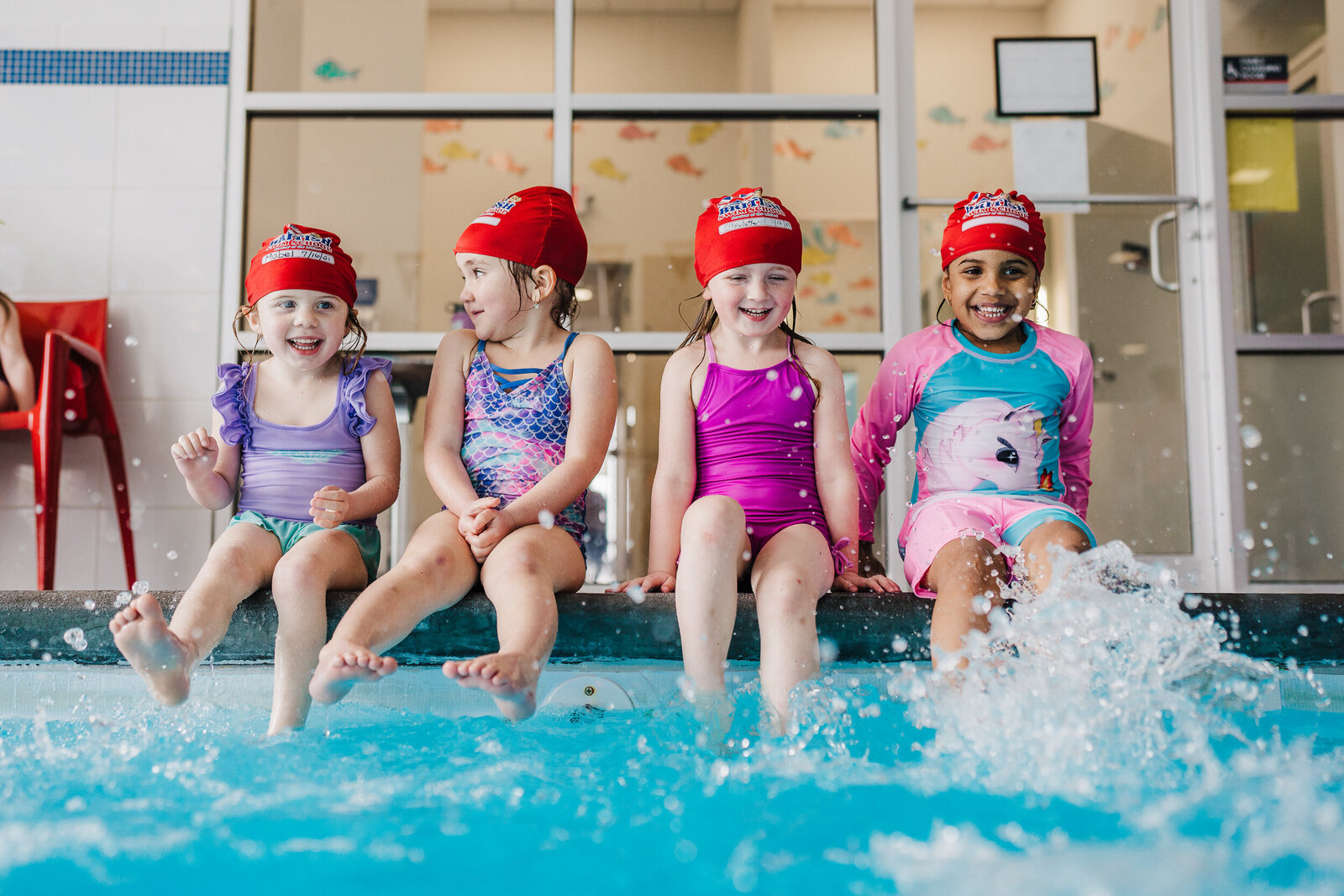 girls with red swim caps sit on side of pool and kick water at camera