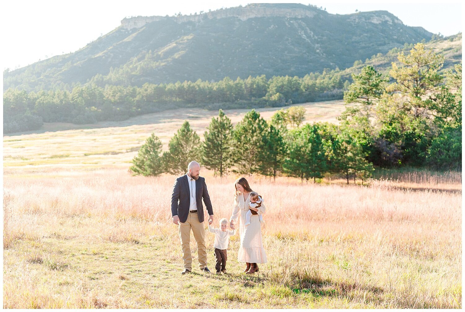 A mom holding a baby and her son's hand walk with dad hold the son's other hand through a field with mountains in the background captured by Denver family photographer