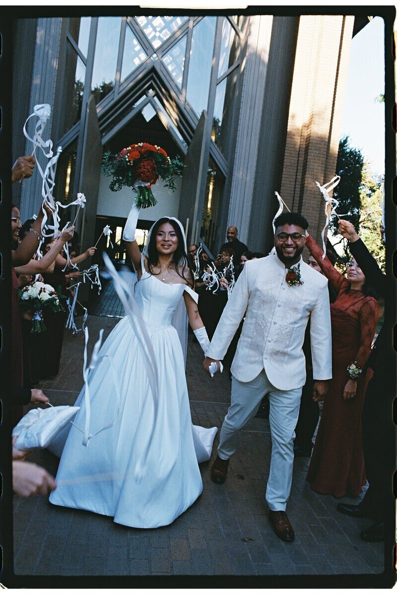 A bride and groom joyfully exit a modern building, holding hands. The bride wears a white dress and holds a bouquet, while the groom is in a suit. Guests cheer and wave white streamers around them in celebration.