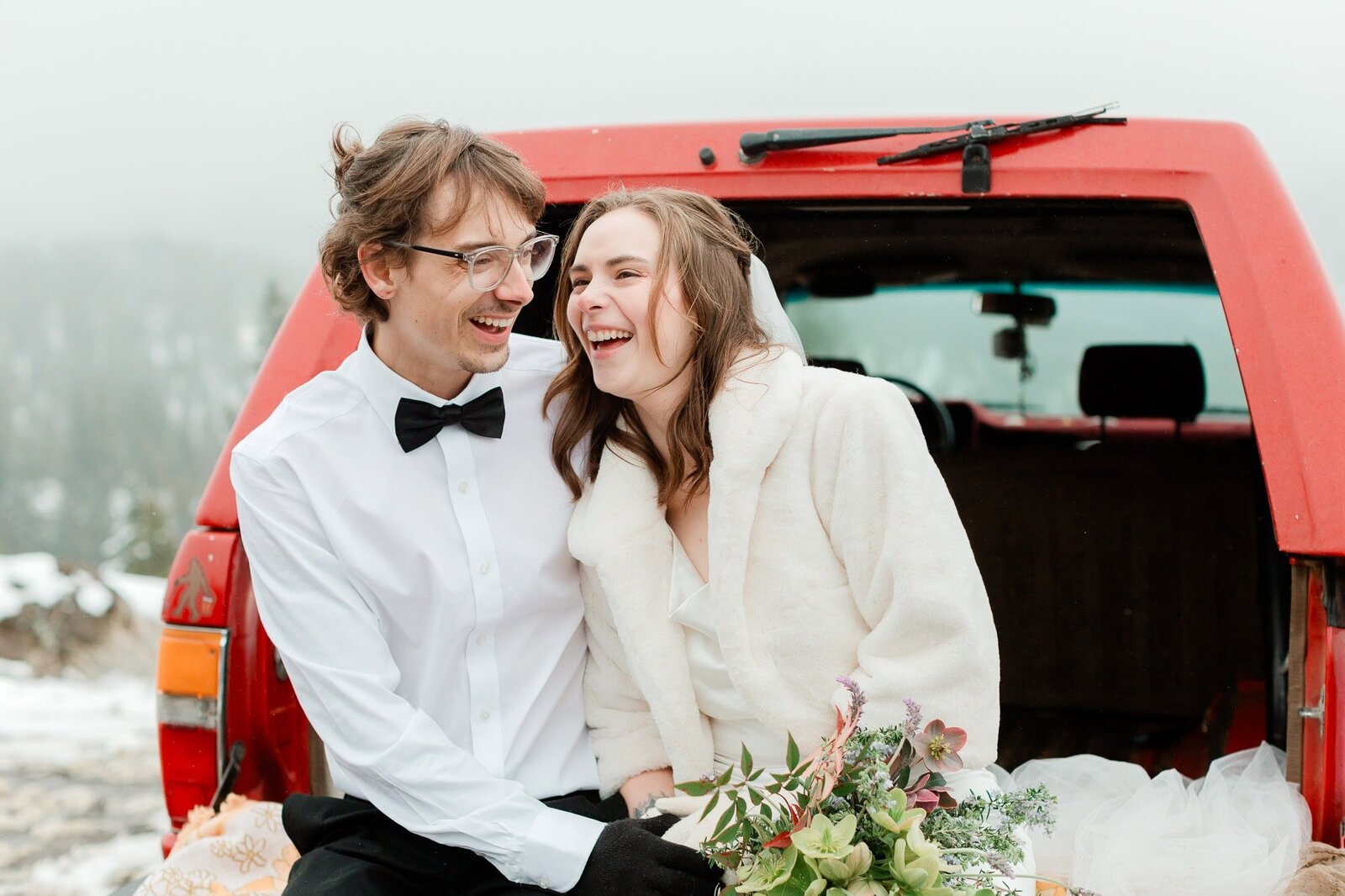 A bride and groom laugh together from the back of their Toyota tailgate while on their winter mountain wedding. 