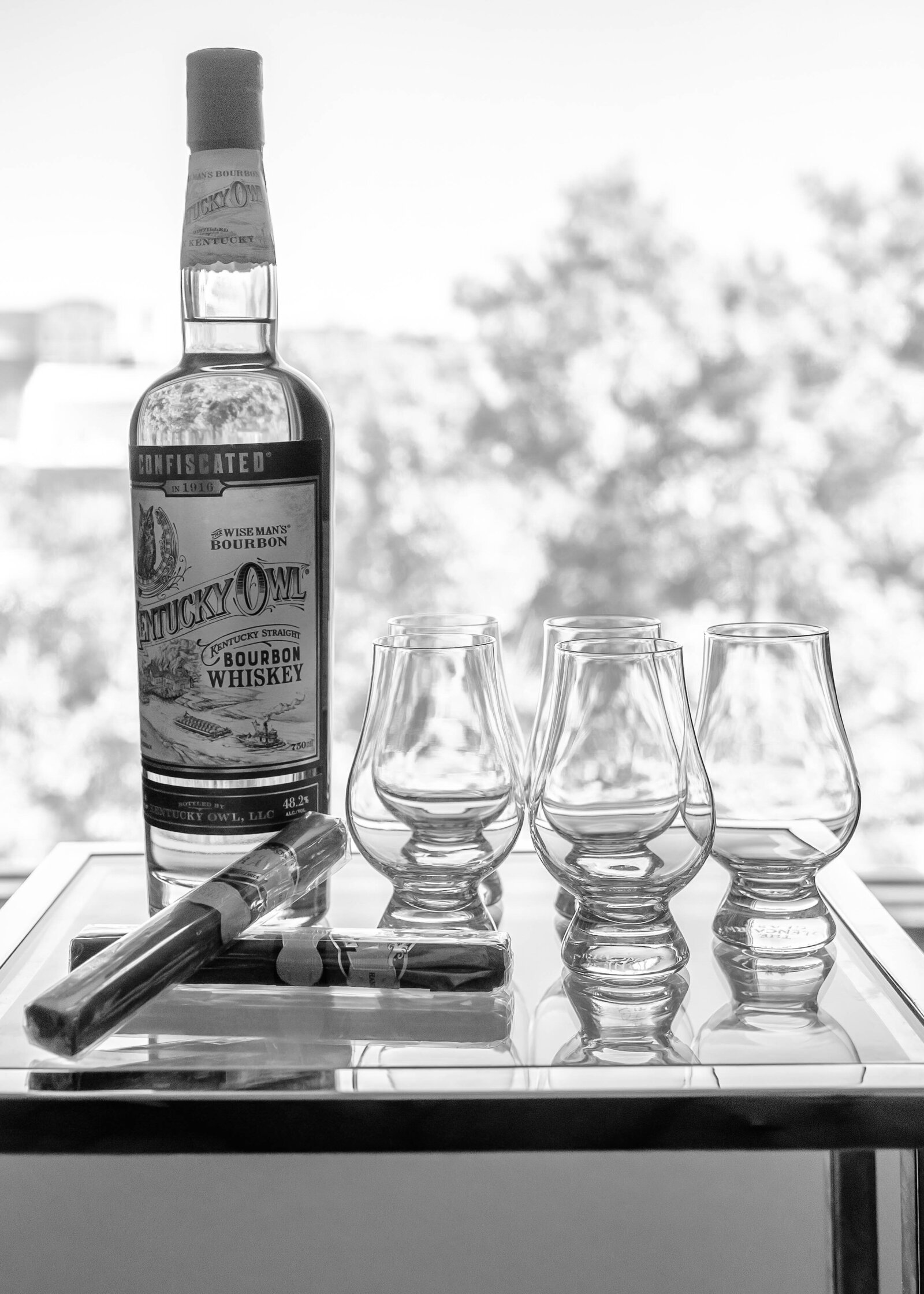 Black-and-white image of a bottle of bourbon, cigars, and glassware arranged on a mirrored table, captured as part of the groom’s wedding day details at a Wilmington, NC wedding, showcasing rustic sophistication.