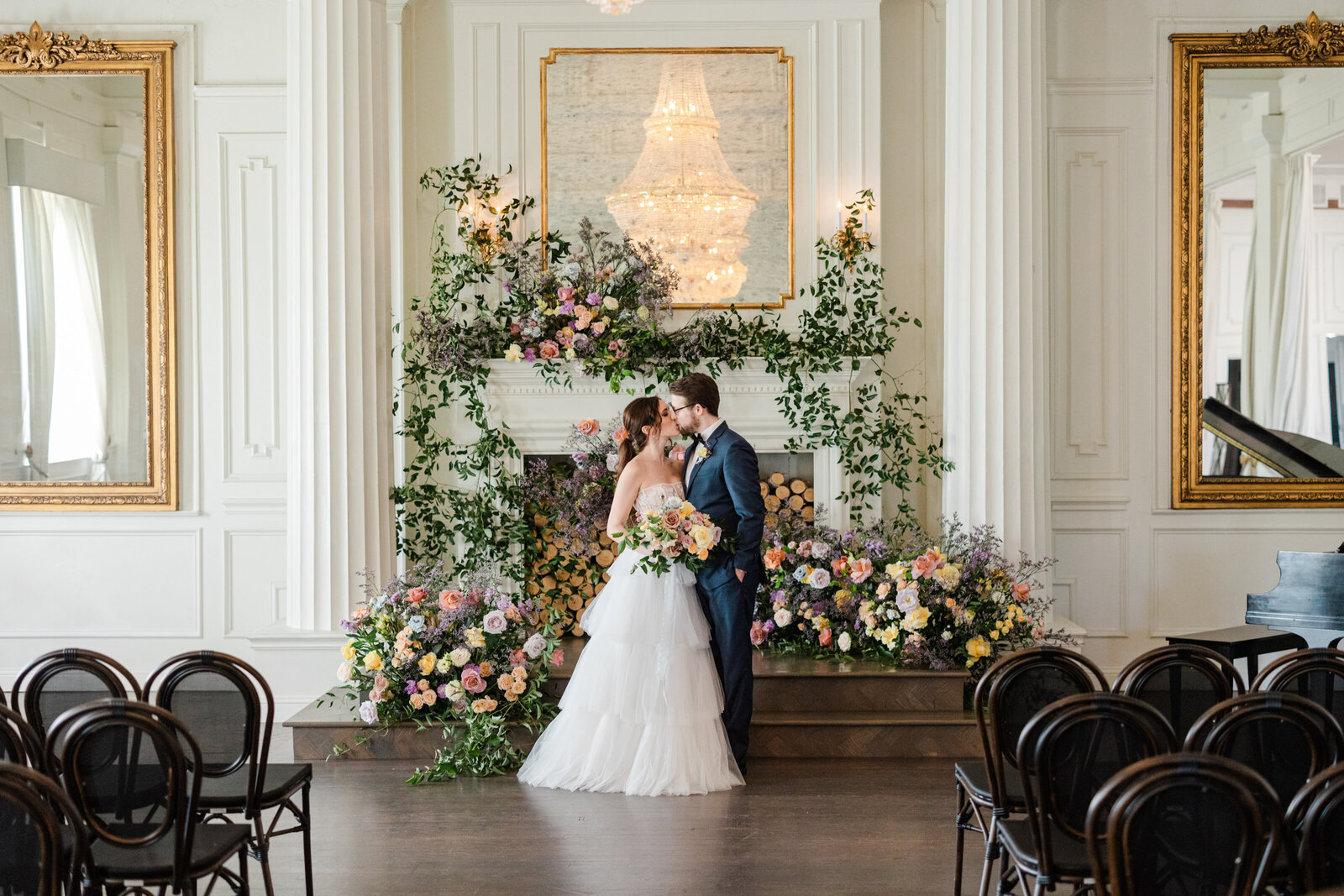 A newlywed couple stands in front of a fireplace at The Mason Wedding Venue in Dallas, TX. They are surrounded by greenery and bold pink, orange, purple, and yellow spring florals. The bride is on the left and is wearing a ruffled tiered wedding skirt and a sheer lace strapless top. She has brown hair and it's in a party ponytail. She's kissing her groom. The groom is wearing a navy blue suit.