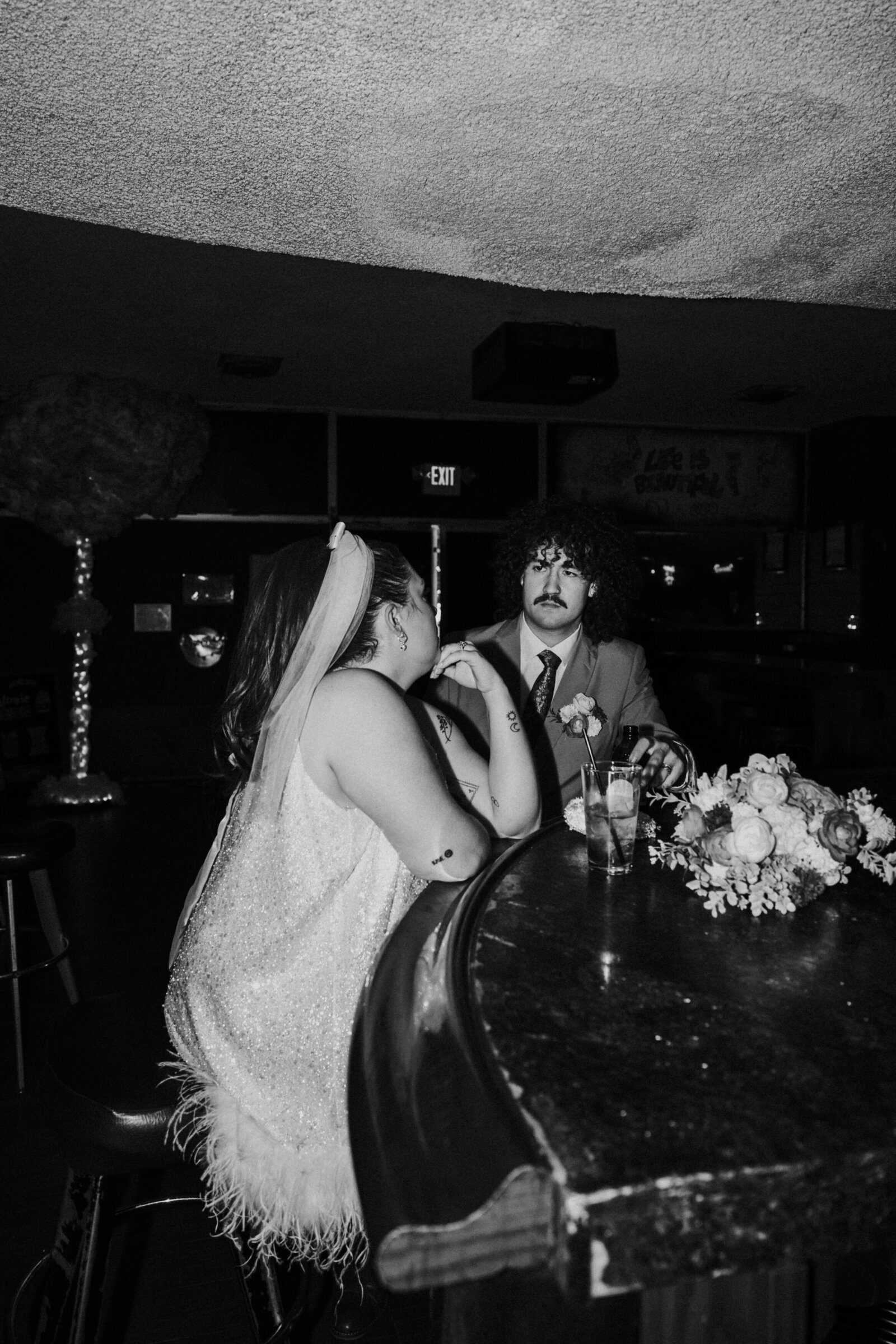 A couple sits at the bar on their elopement day, smiling at one another.