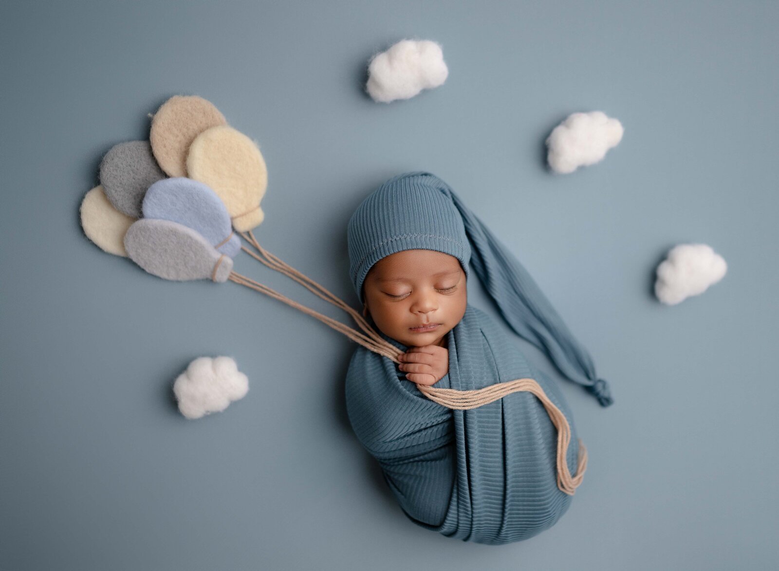 Baby swaddled in blue wrap, holding felted balloons and surrounded by clouds. Newborn Photography session photographed at Jenni Nikki's photo studio in Marietta, GA, near Atlanta.