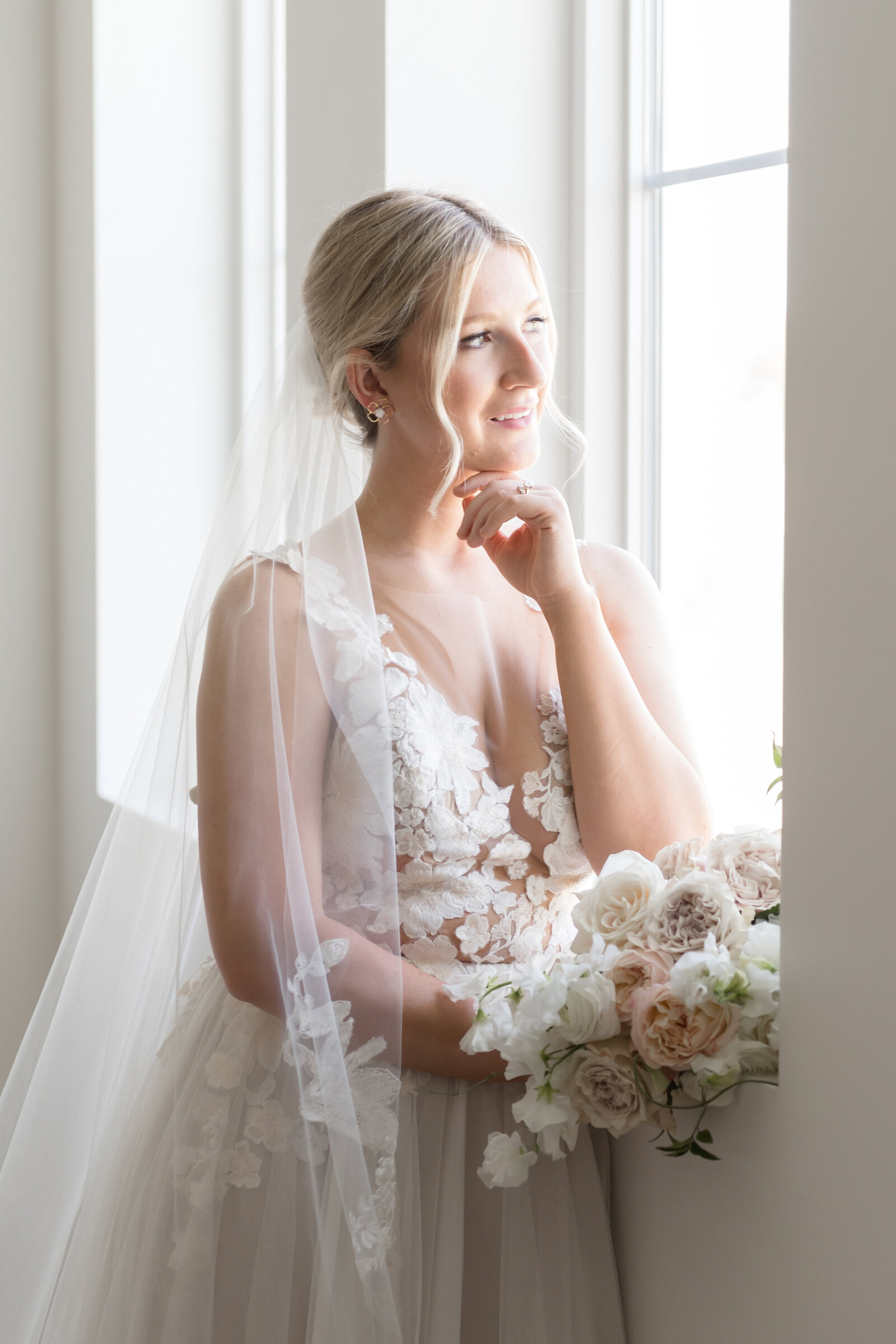 bride standing looking out a window with her hand propped under her chine and her bouquet in her other hand