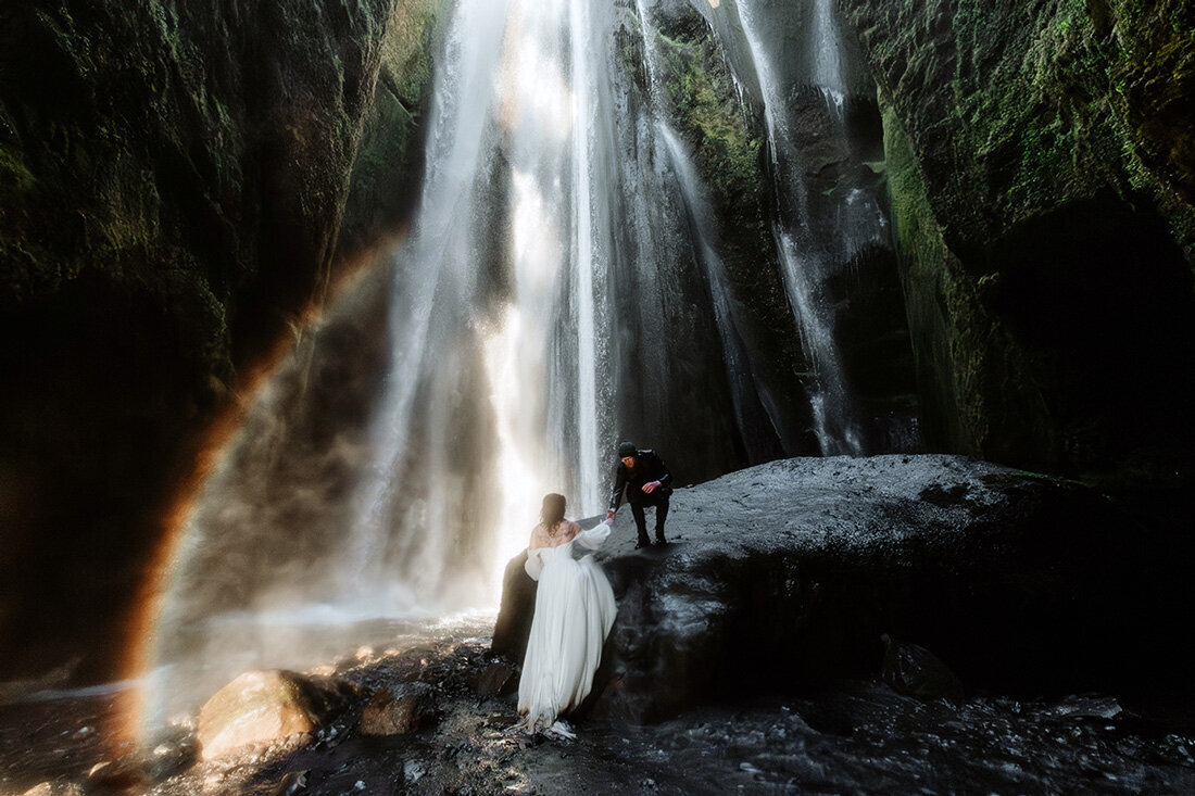 Dramatic photo of a couple climbing up on a slippery stone in a cave that has a waterfall and rainbow in Iceland