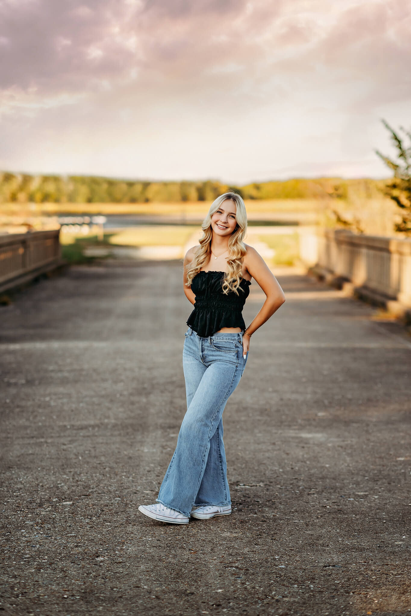 A person with long blonde hair stands smiling on a bridge with a scenic background. They're wearing a black top, blue jeans, and white sneakers, with a colorful sky overhead.