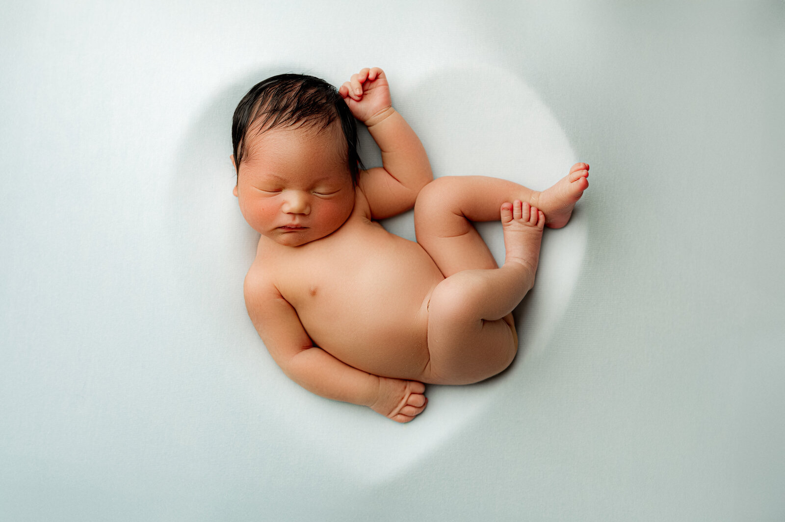 Captured by a Charlotte newborn photographer, this image shows a newborn baby peacefully sleeping on a white blanket. The dark-haired infant lies curled, hand on chest, surrounded by the blankets subtle heart shape—a serene moment expertly documented.