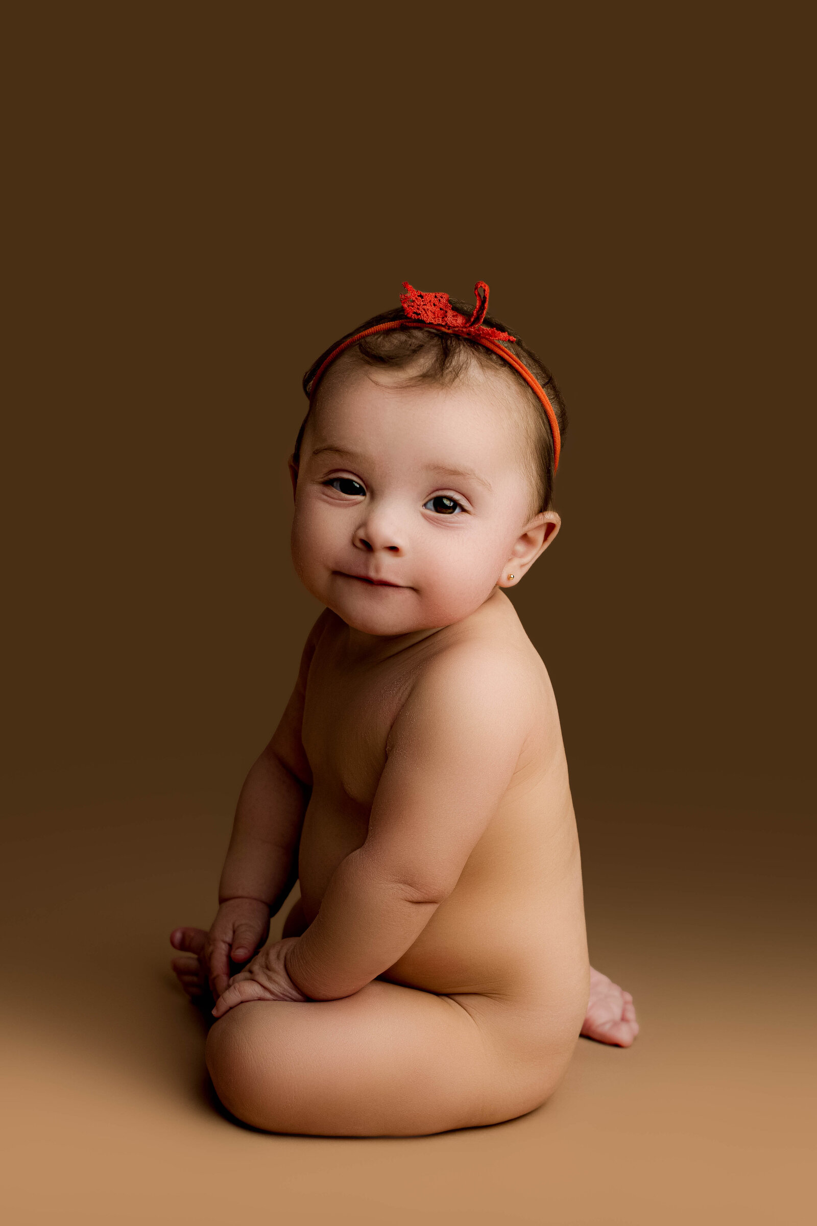 Bare baby girl with an orange headband sitting up confidently with little smirk on a rich brown backdrop for her Charlotte milestone portrait.