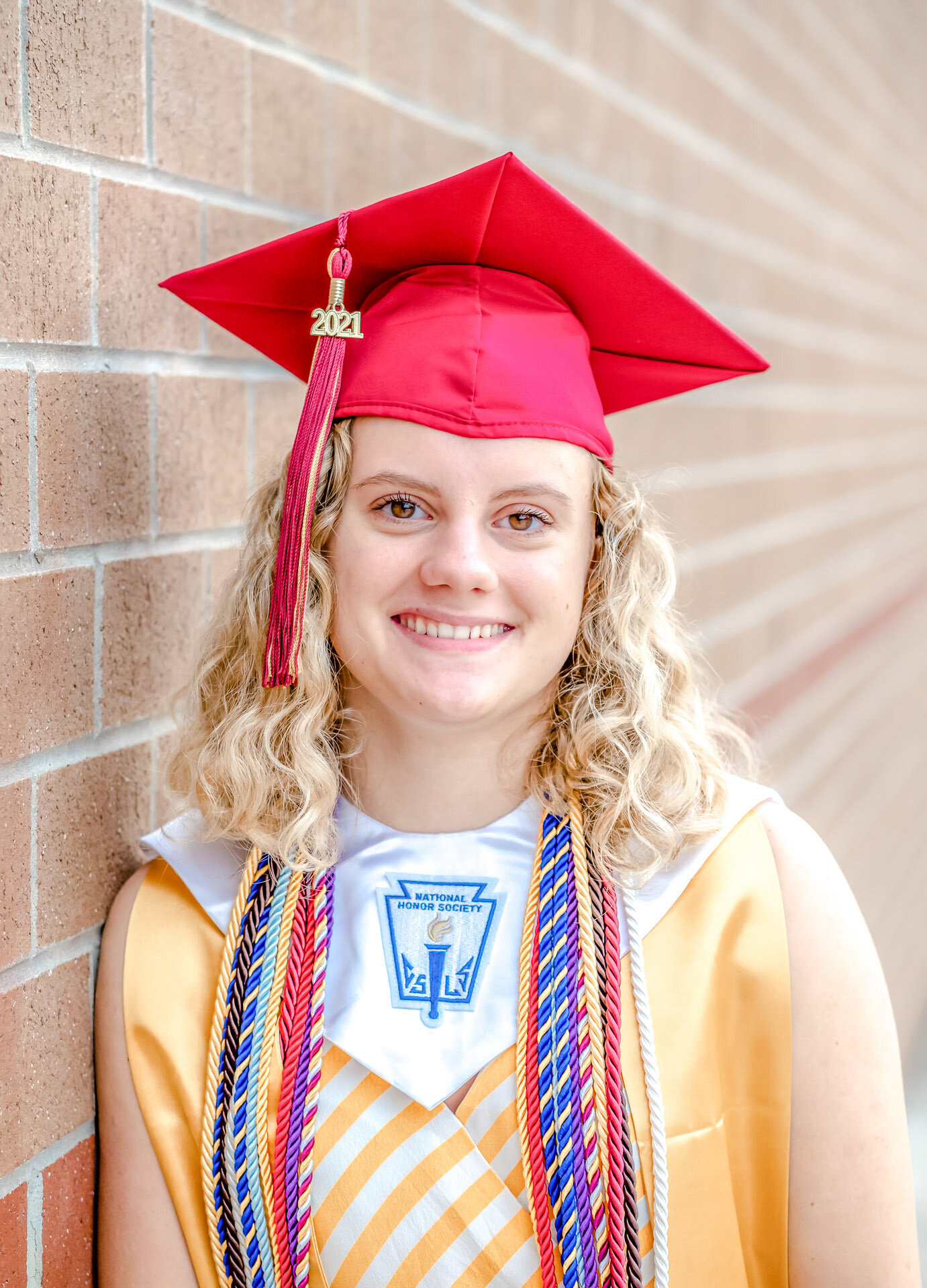 An Ashley High School senior in a red graduation cap and honors cords leaning against a brick wall in Wilmington, North Carolina. This portrait captures academic achievement, perfect for those seeking professional senior photography.