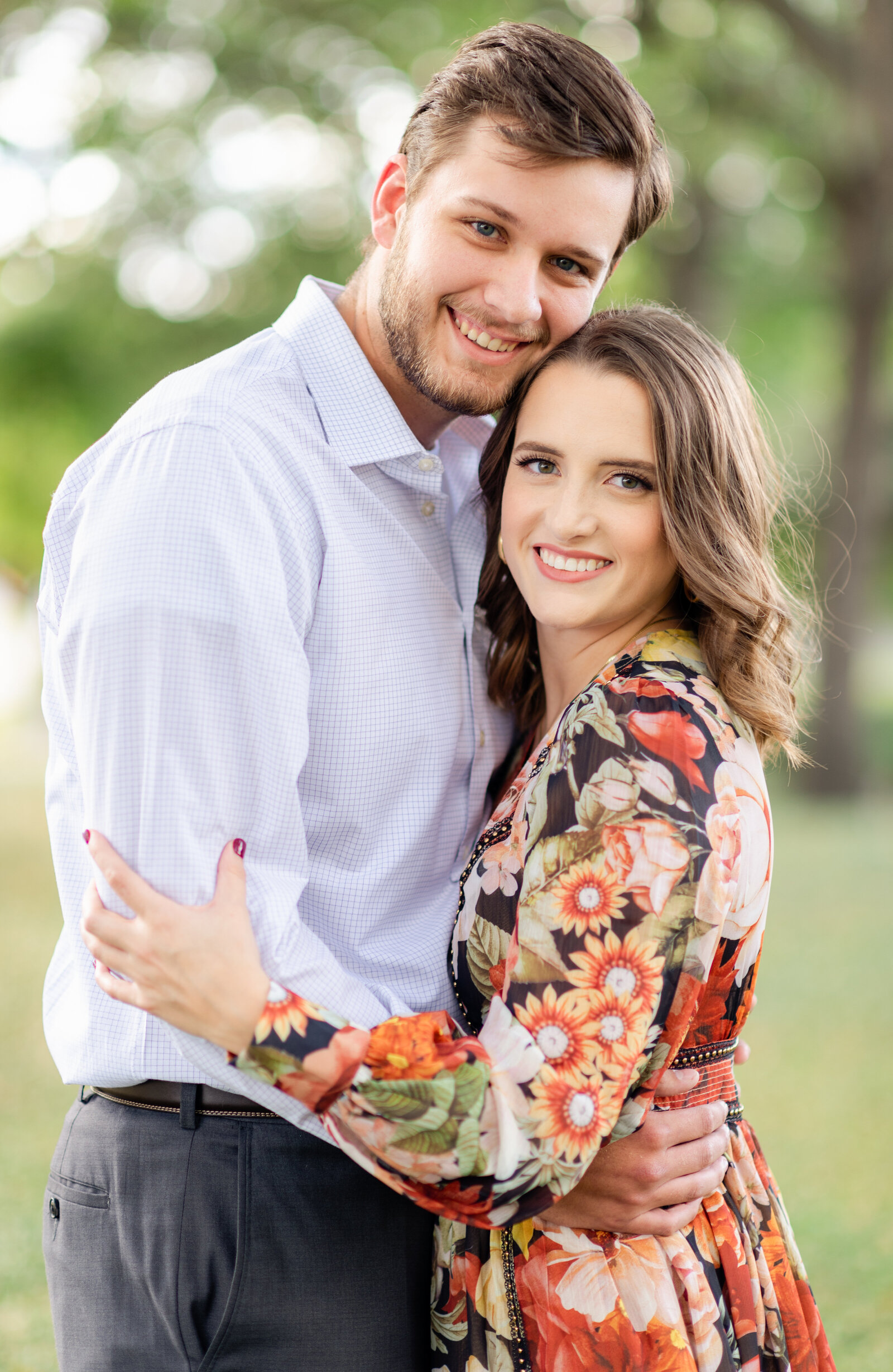 Portrait of a man in a white dress shirt and gray slacks holding a woman wearing a floral dress while smiling  outdoors.