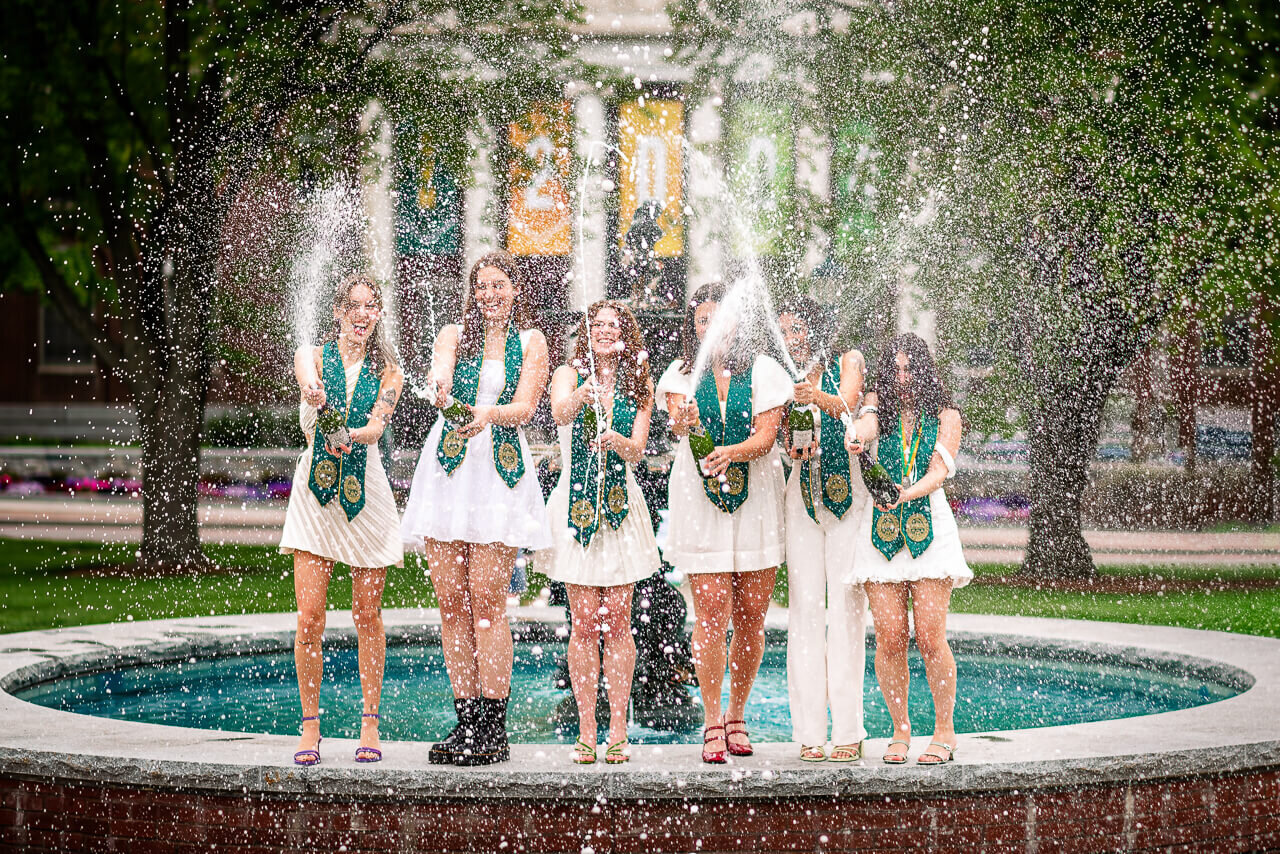 group of young women celebrates their University of Vermont graduation by spraying champagne in front of the UVM fountain in Burlington, Vermont, filled with joy and excitement.