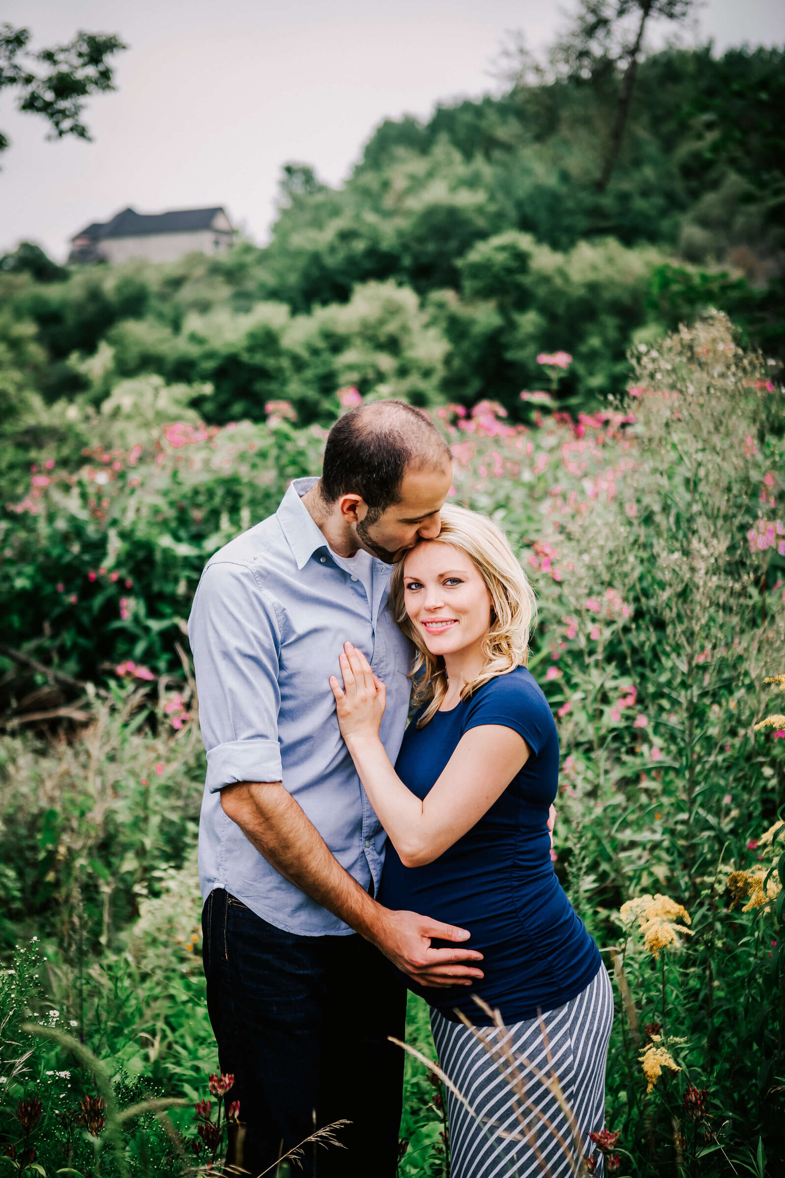 husband and wife stand in a flower field while expecting their first baby