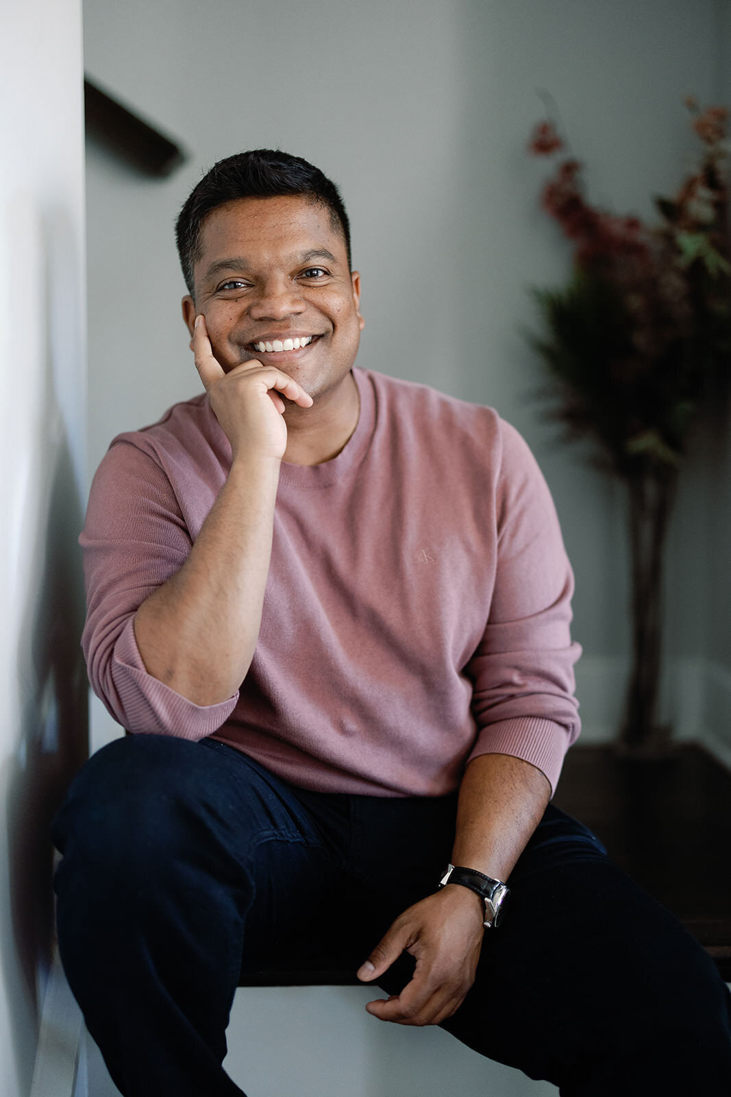 Professional portrait of a smiling man in a relaxed pose, wearing a casual pink sweater in a modern home.