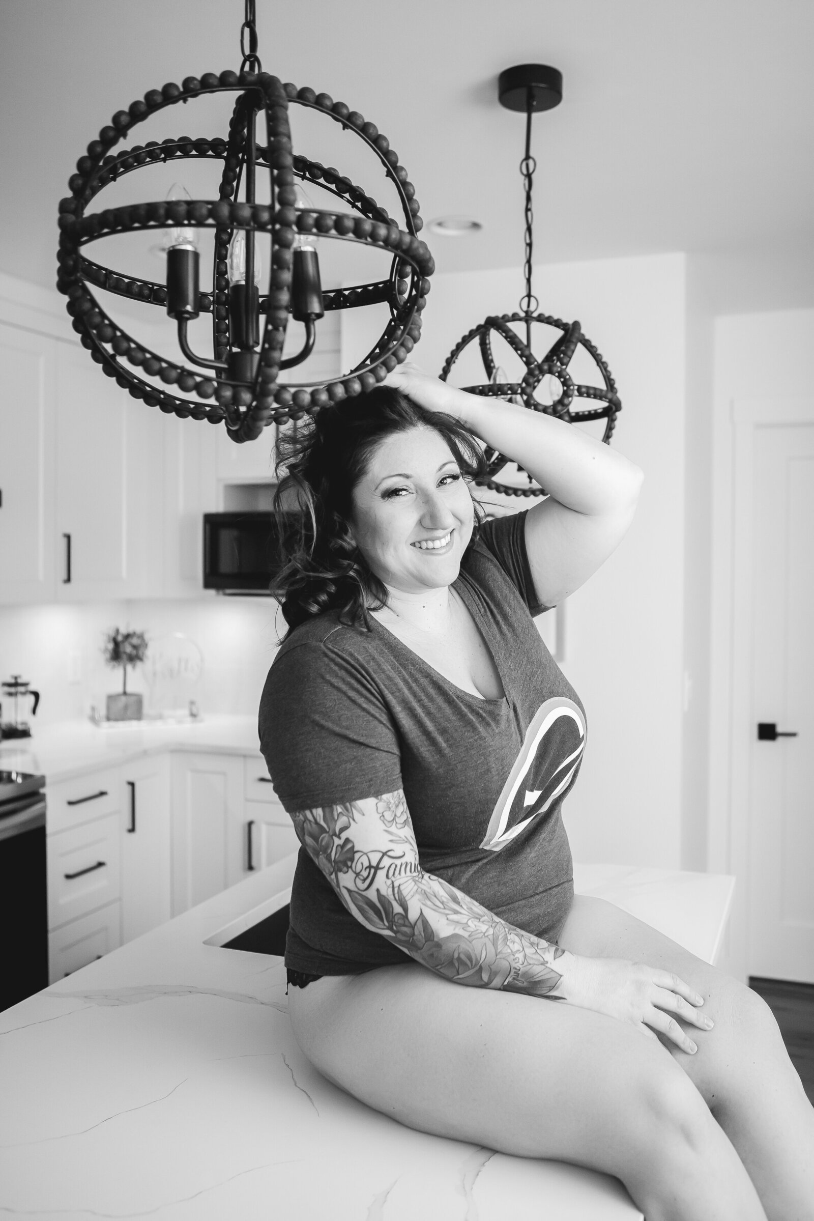 A black-and-white image of a smiling woman sitting on a kitchen counter, showing off her tattoos and laid-back style.