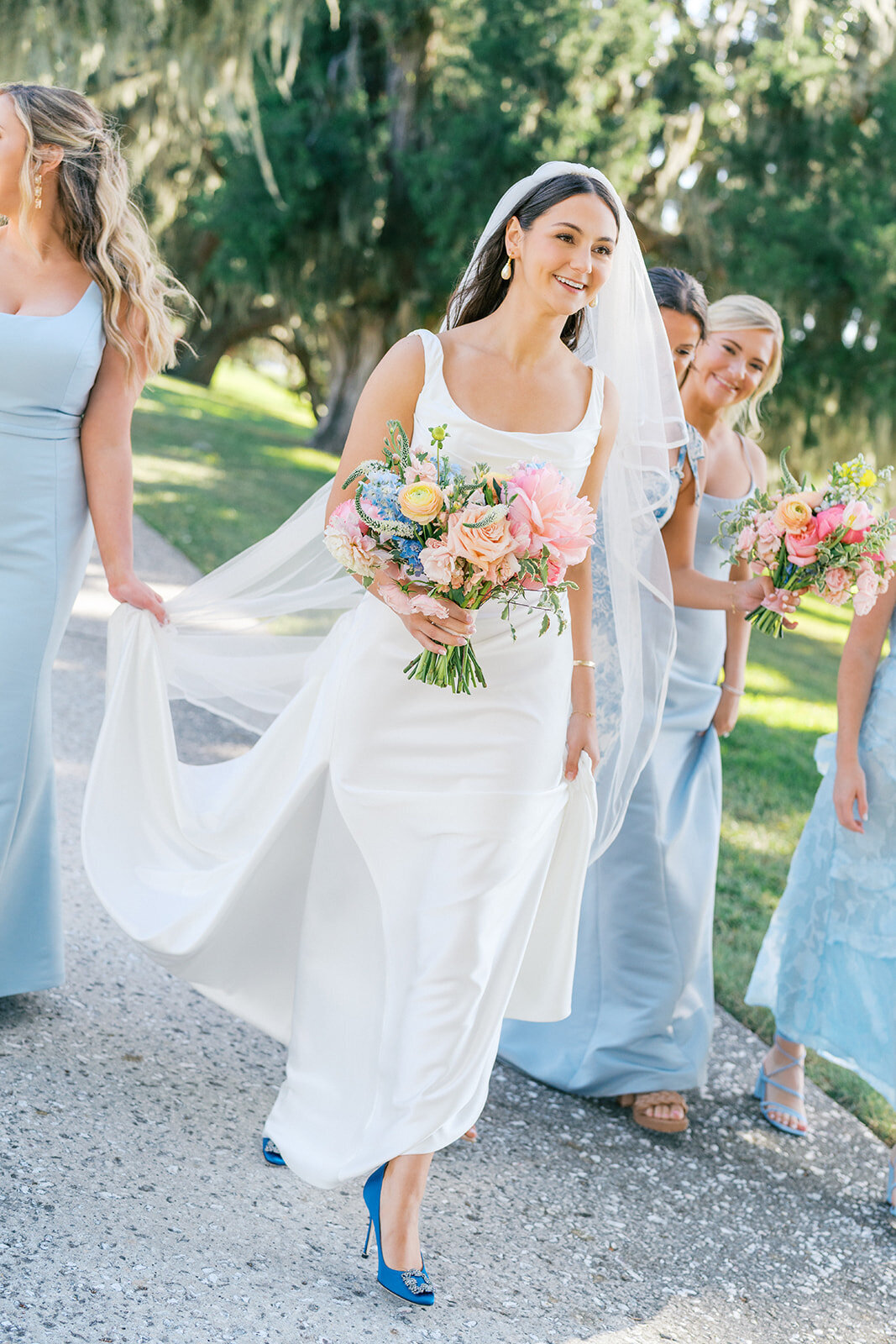 Bridesmaids in light blue hold bride's dress and veil walking down walkway art Jekyll Island Club Resort