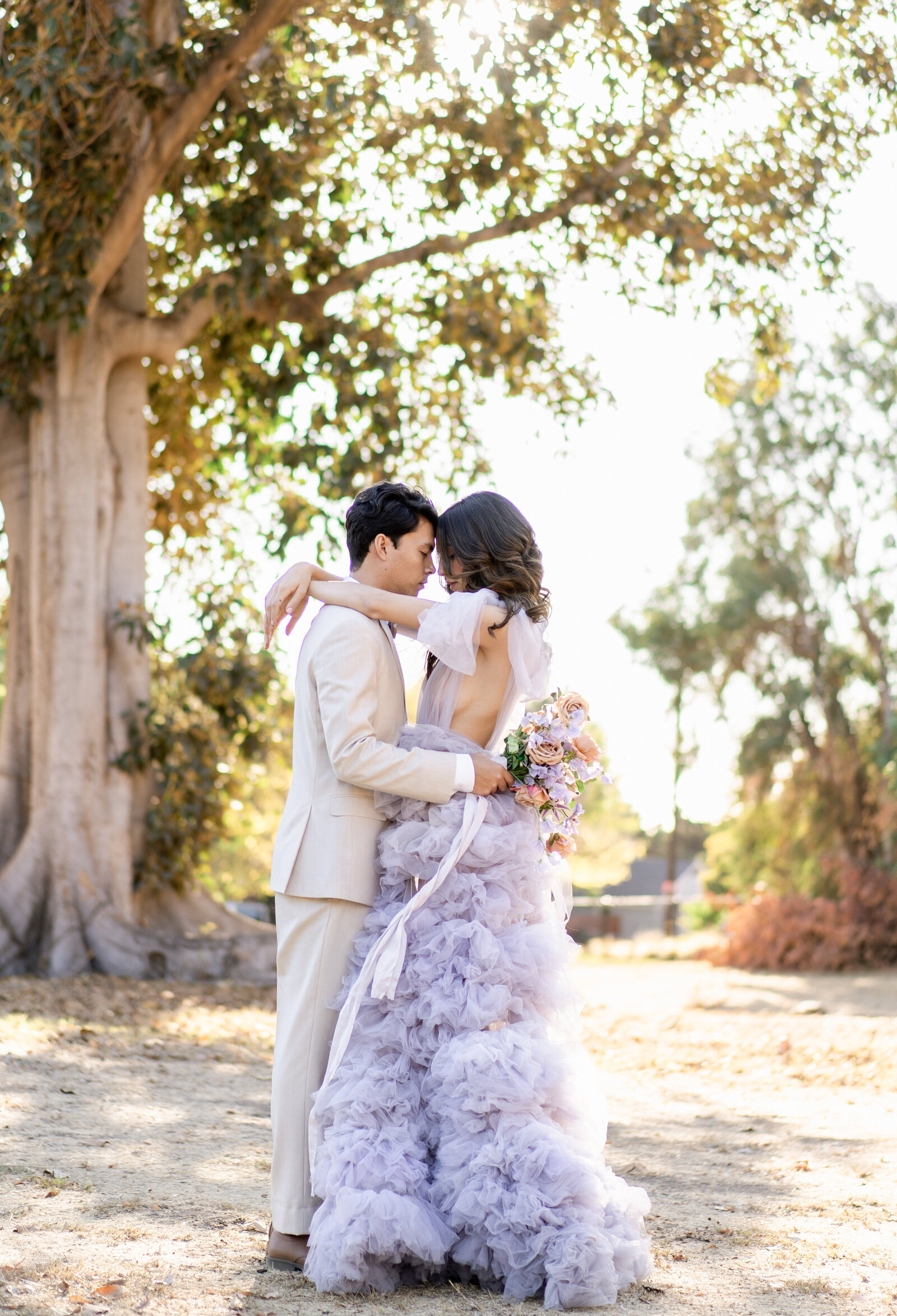 Bride and groom in a lavender gown and cream suit hold each other outdoors in front of large tree