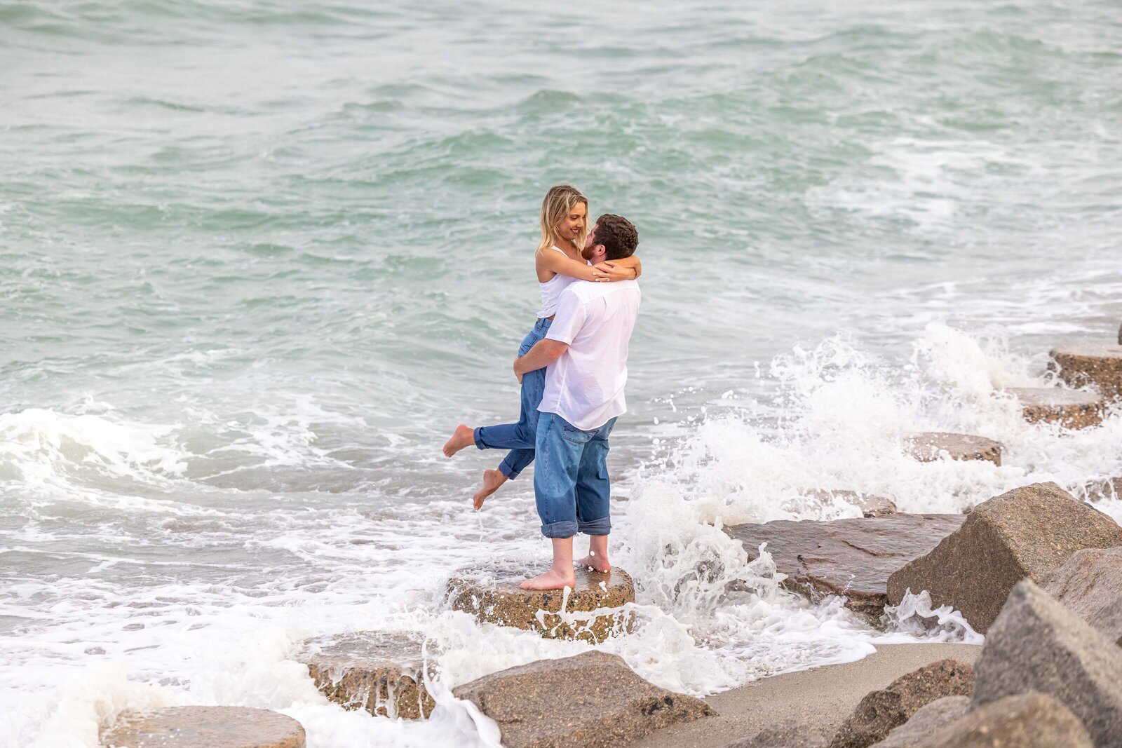 An engaged couple embracing on rocks at Fort Fisher as ocean waves crash around them in North Carolina. This dynamic and romantic photo captures the thrill of artistic wedding photography, appealing to couples seeking adventurous engagement photos.