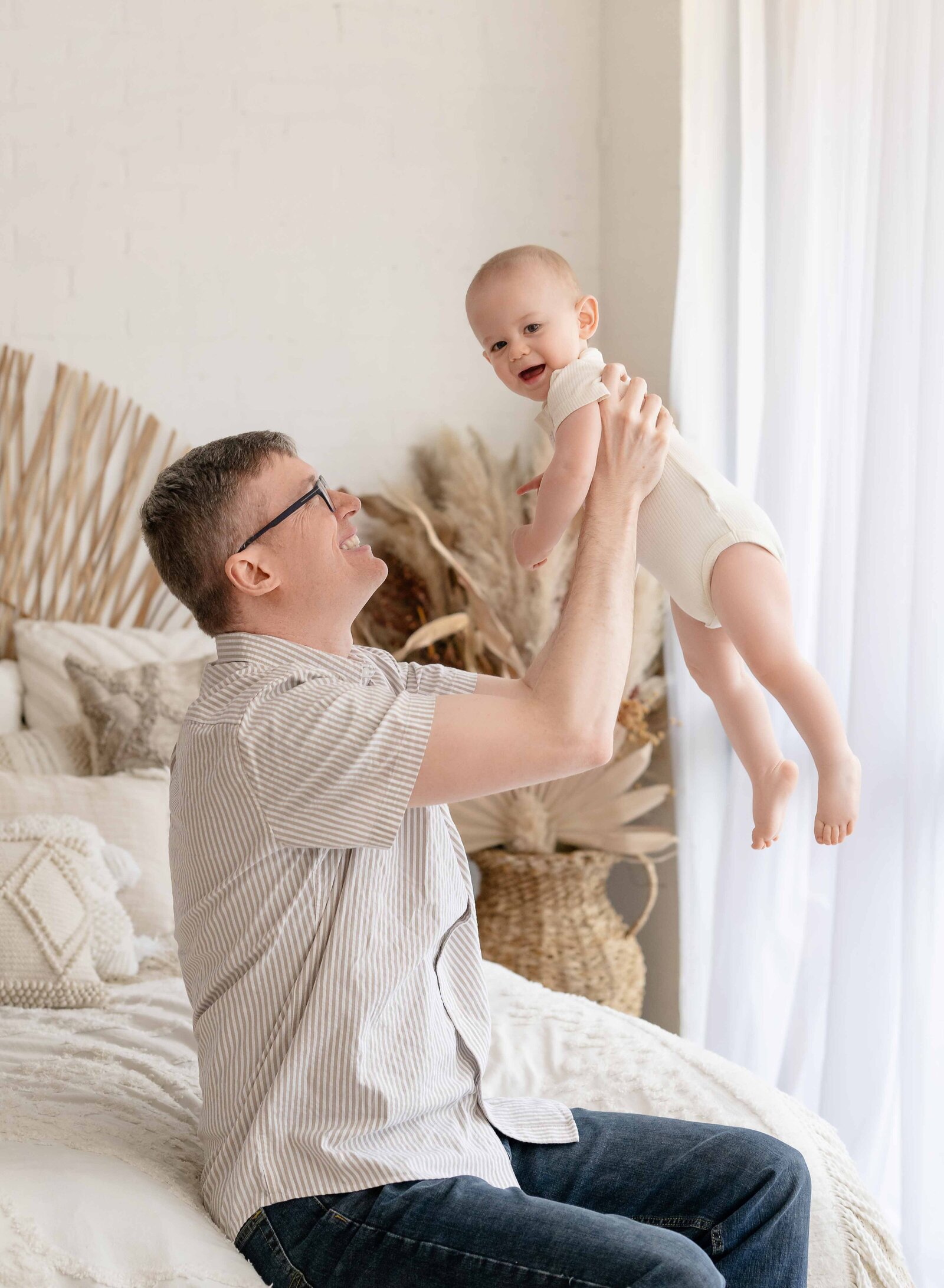 Dad sitting on bed and holding baby up in the air. Photographed in a lifestyle bedroom at Atlanta family photo studio.