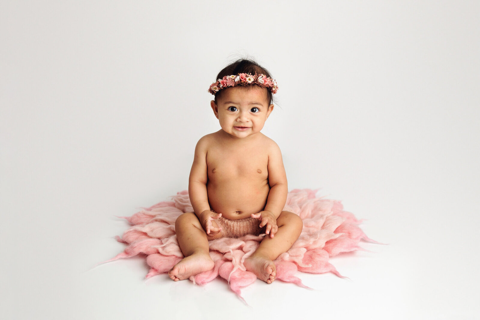 An adorable baby with dark hair and big eyes sits on a soft, pink petal-shaped blanket against a white background. The baby wears a floral headband and a light pink diaper cover, smiling sweetly at the camera. The setting is bright and airy, highlighting the baby's joyful expression and the delicate details of the blanket and headband.