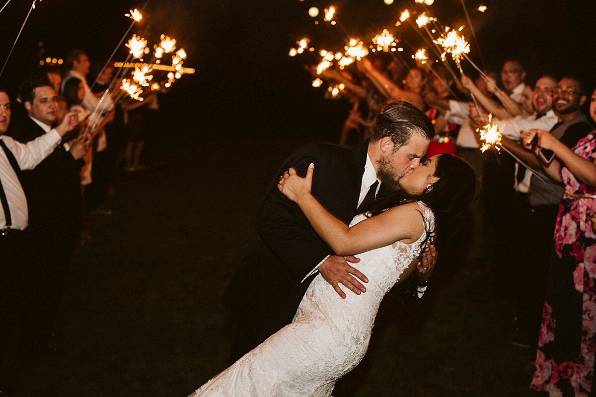 Groom dips the bride into a kiss as they leave their wedding to a sparkler send off