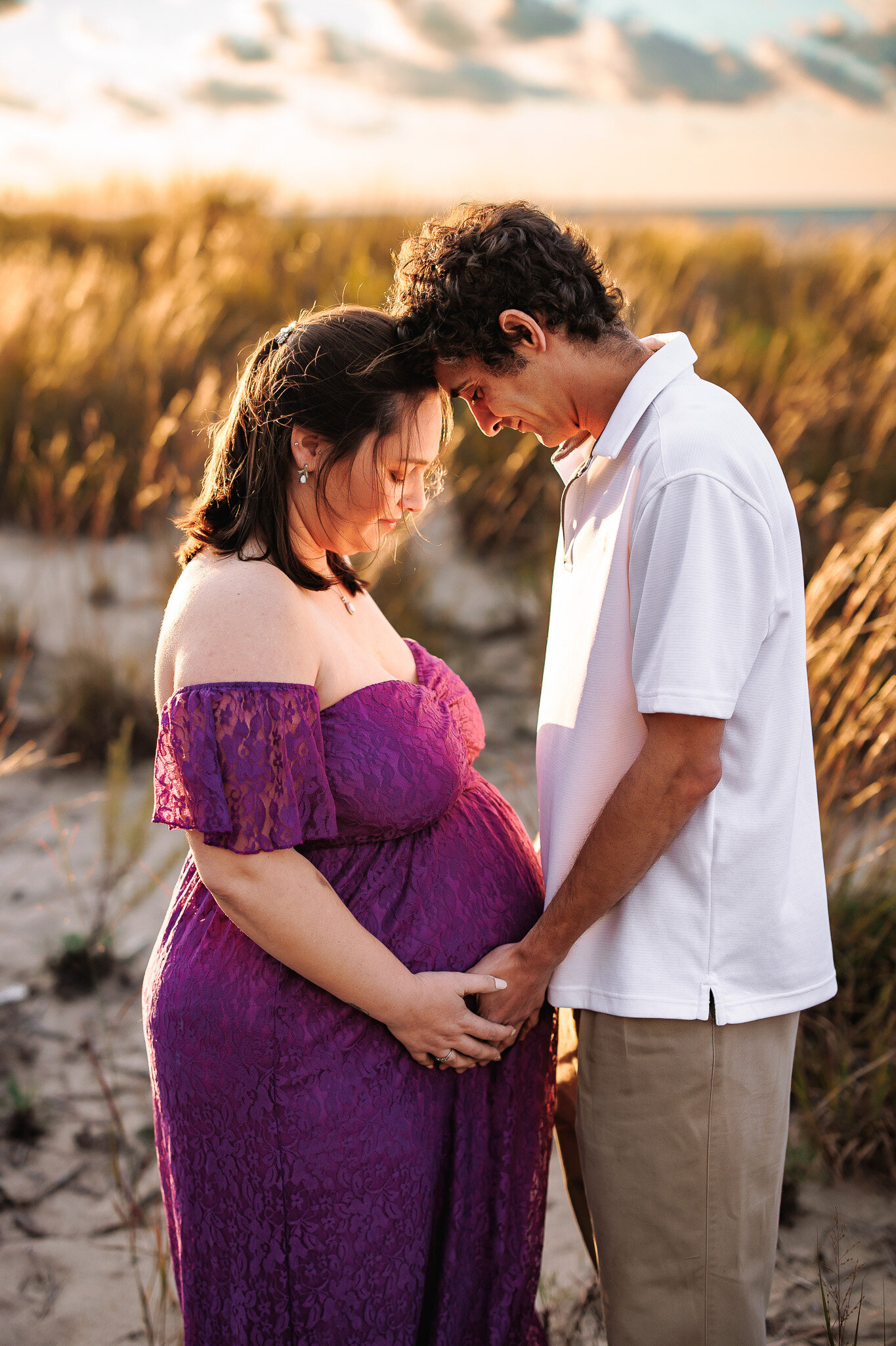 Foreheads together, an expecting couple look down at their baby to be amongst the golden beach grass