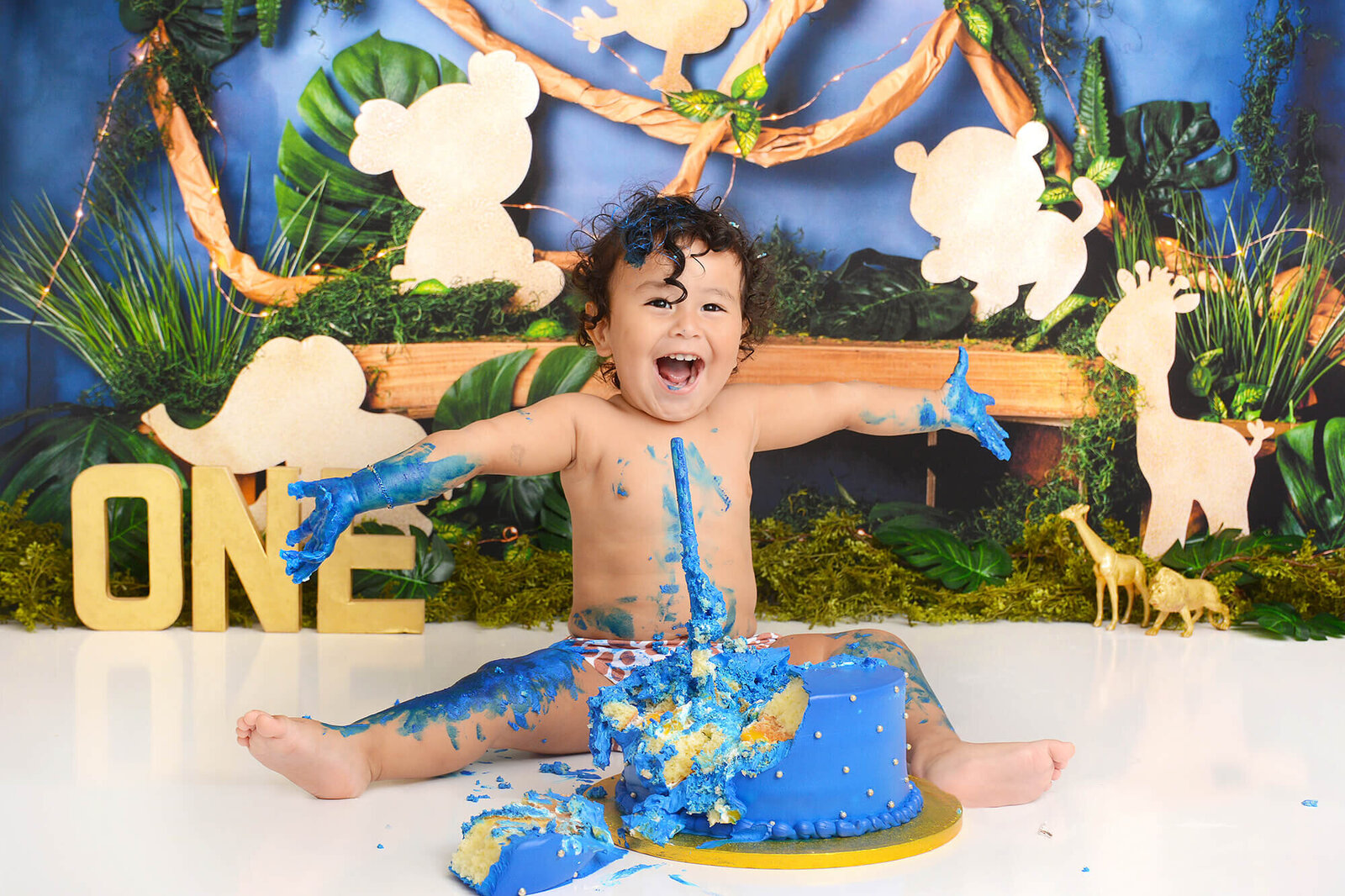 birthday boy covered in blue frosting from his birthday cake smiling in front of a jungle background