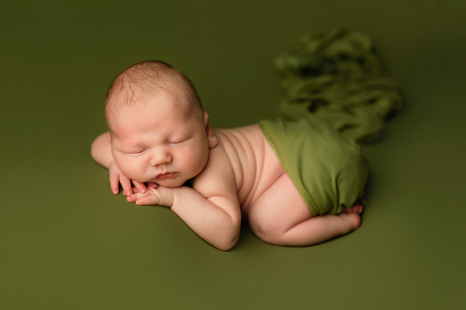 Baby boy posed on belly with chin resting on hands in front of an olive green backdrop 