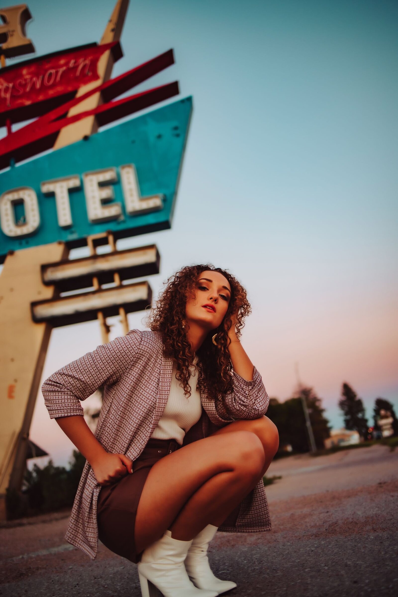 a young girl in white boots, a Burgundy skit, a light  purple jacket and white shirt posing in a squatting position in front of a motel sign in Nebraska.
