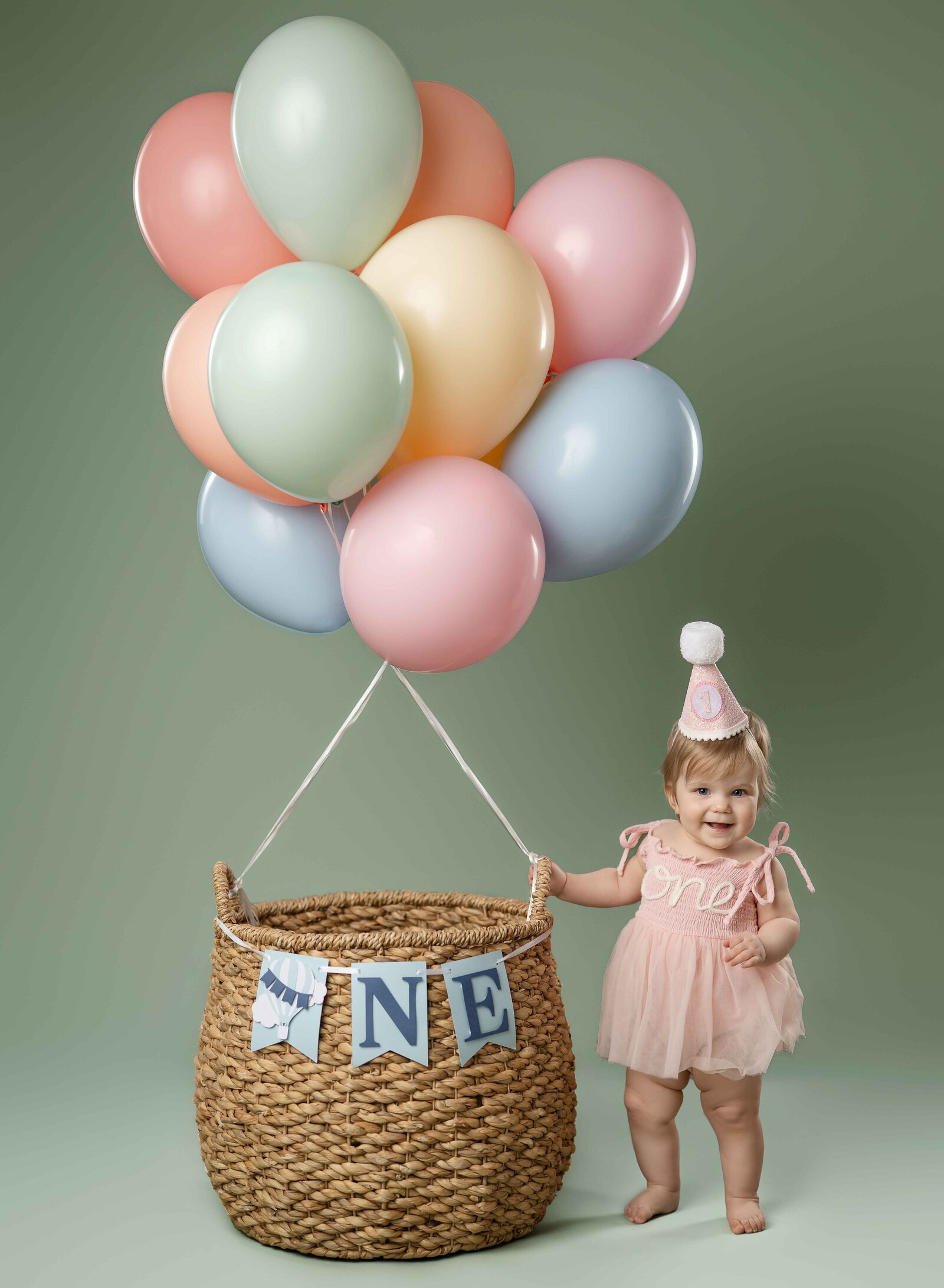 Baby girl in pink ONE dress standing next to pastel balloon basket and smiling for 1st birthday photo at Atlanta photo studio.
