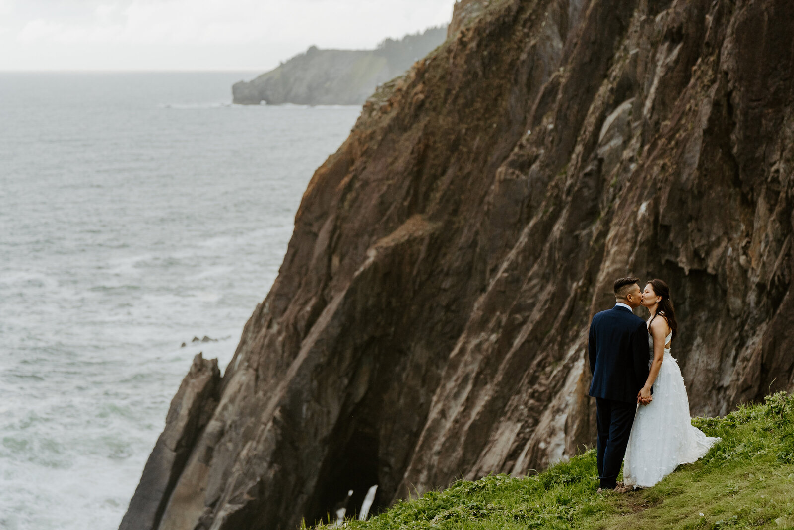 Bride and groom hugging in wedding attire on a grassy hillside with sheer rock cliffs and the Pacific Ocean in the background