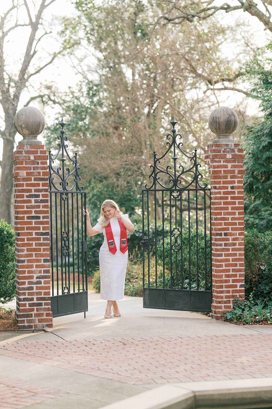 Girl looks down shoulder with red graduation sash in between iron gates and brick pillars on campus