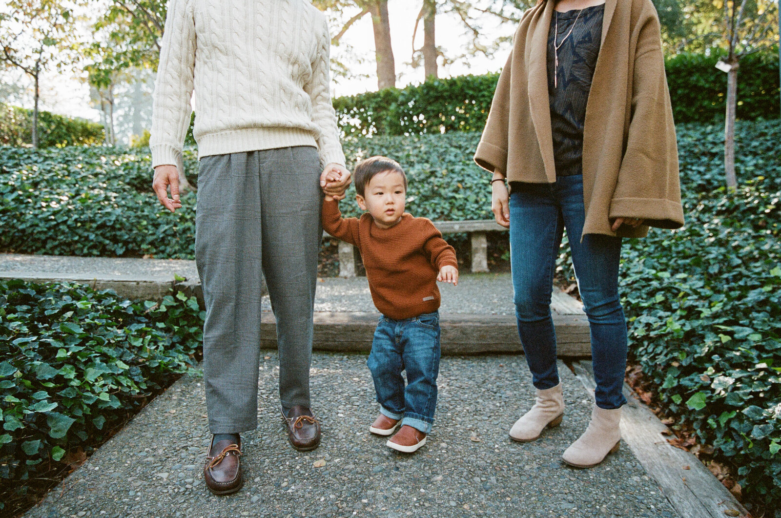 photo of toddler boy in brown sweater and jeans standing between parents and holding dad's hand