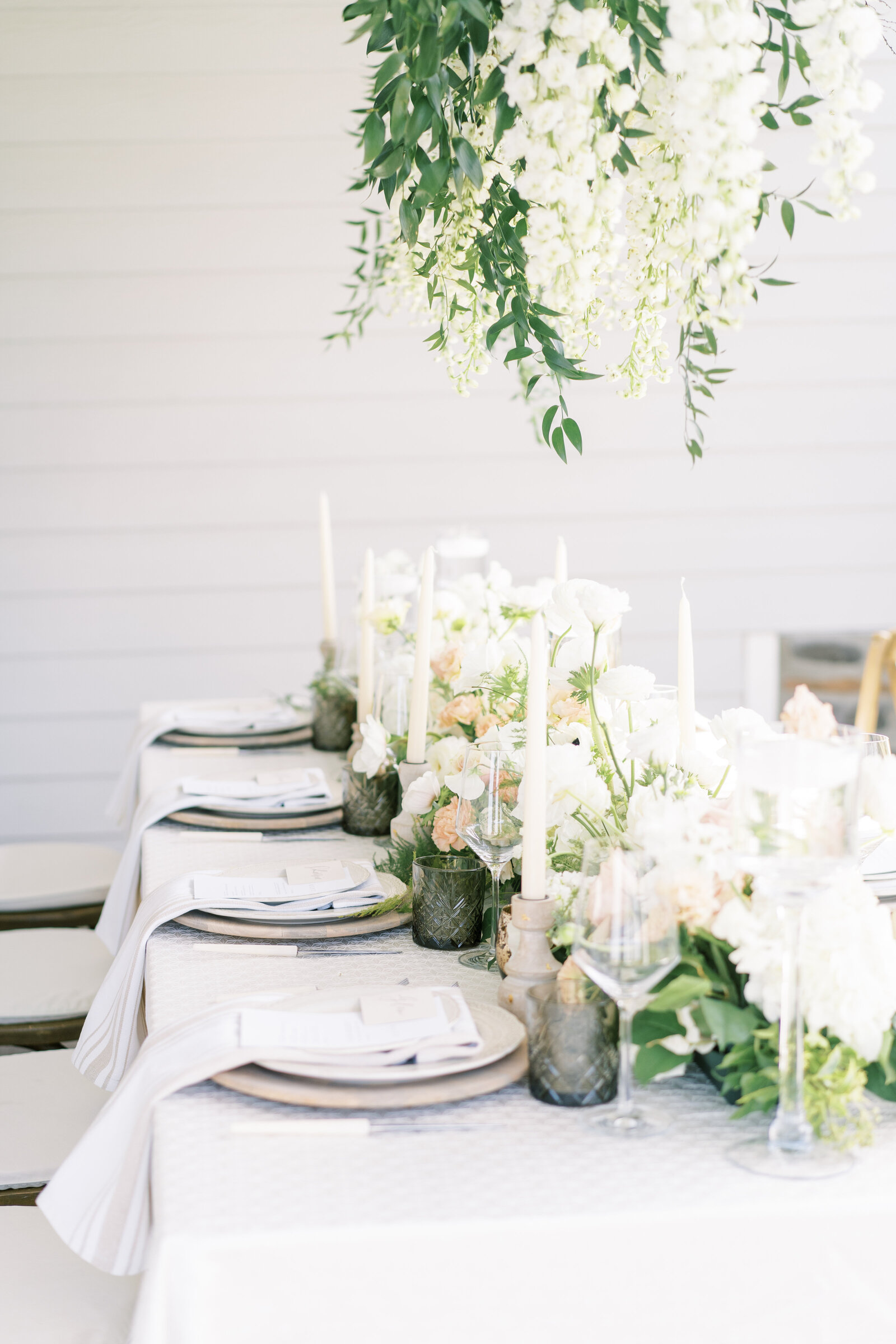 Ivory places, cream candlesticks, and green glassware atop a table dressed in white linens with a hanging centerpiece above.