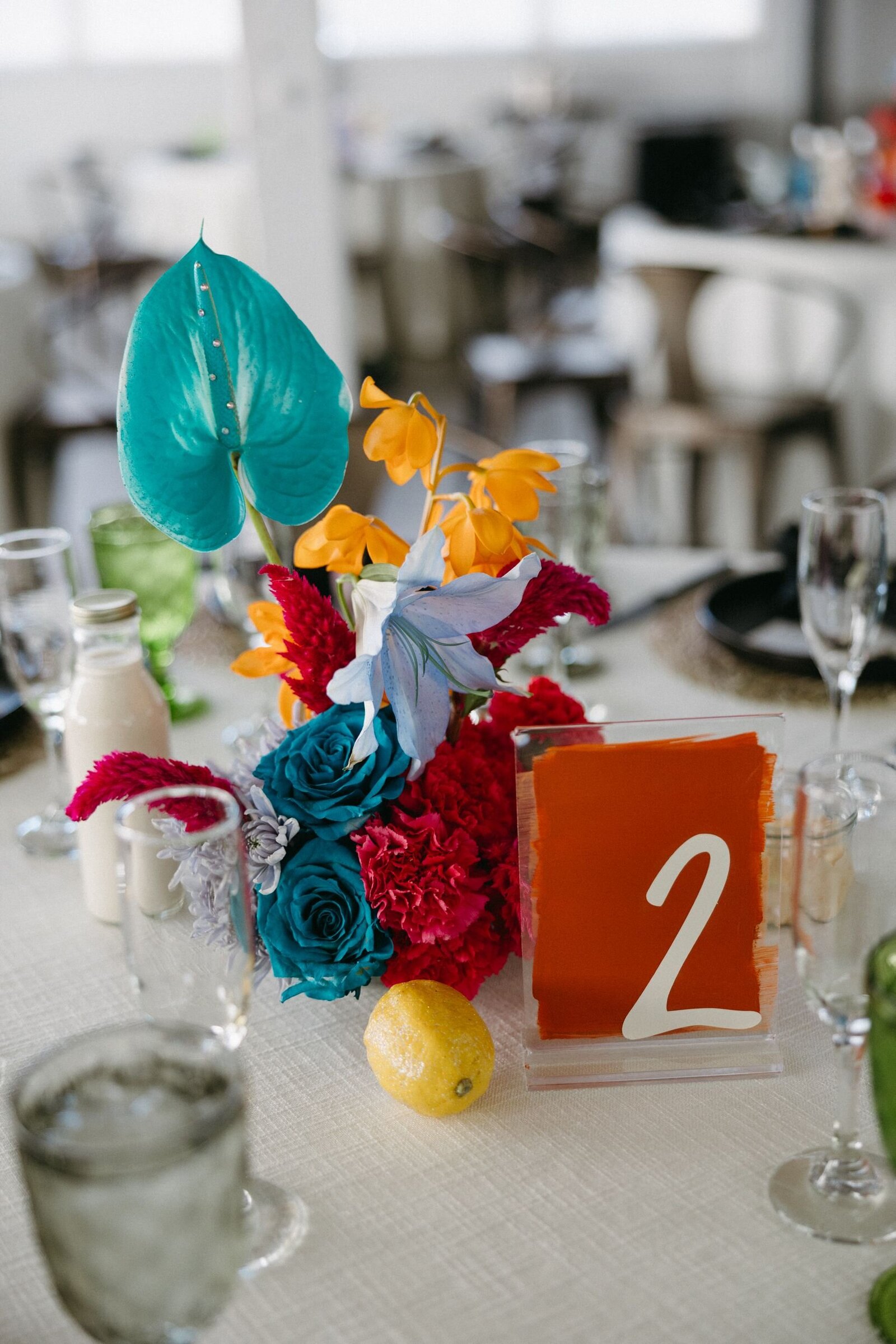 A colorful floral arrangement sits in the middle of a white wedding table with glittery lemons and a bright orange table number