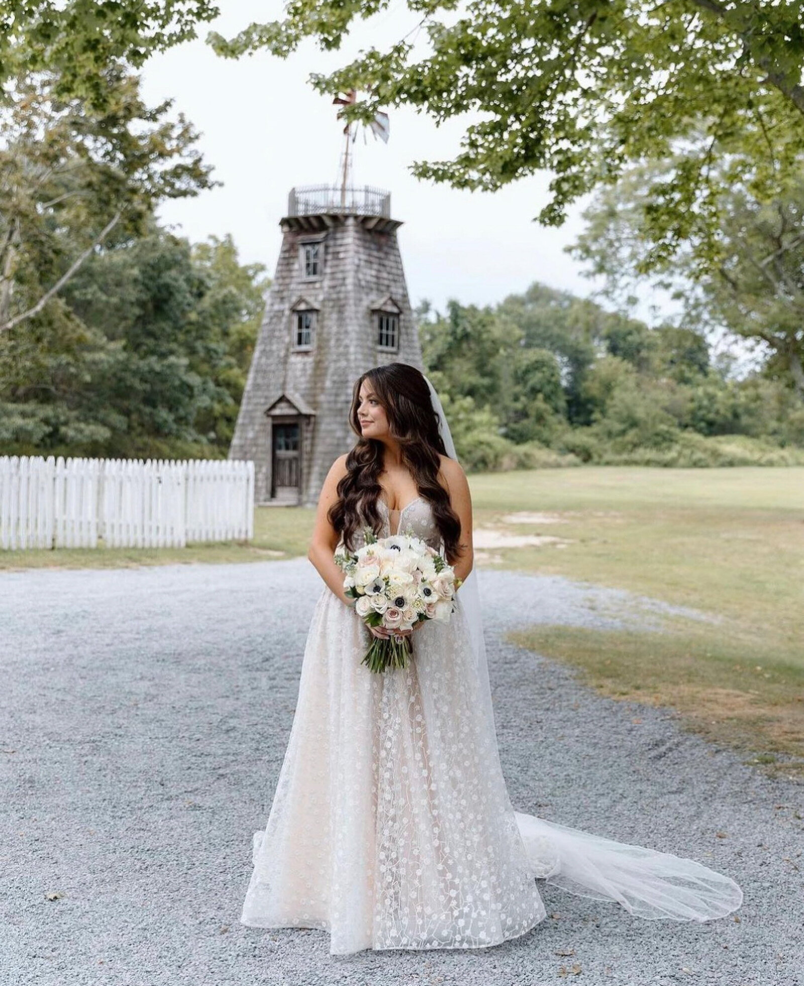bride dressed in a white, lacey wedding gown with a train, and long, wavy hair cascading over her shoulders standing outdoors in front of a rustic wooden windmill, holding a large bouquet of flowers