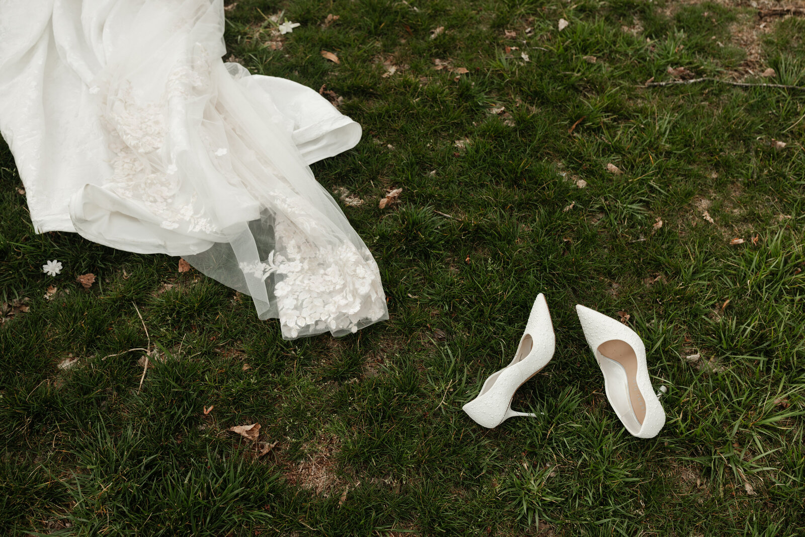 Editorial wedding detail photo featuring a floral lace bridal gown and white heels laid out on the grass, styled with natural textures and romantic light.