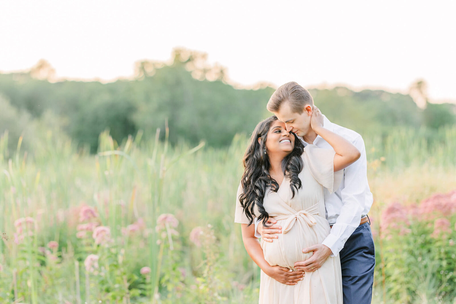 Man embraces his pregnant wife from behind as she reaches up to place her hand on his neck